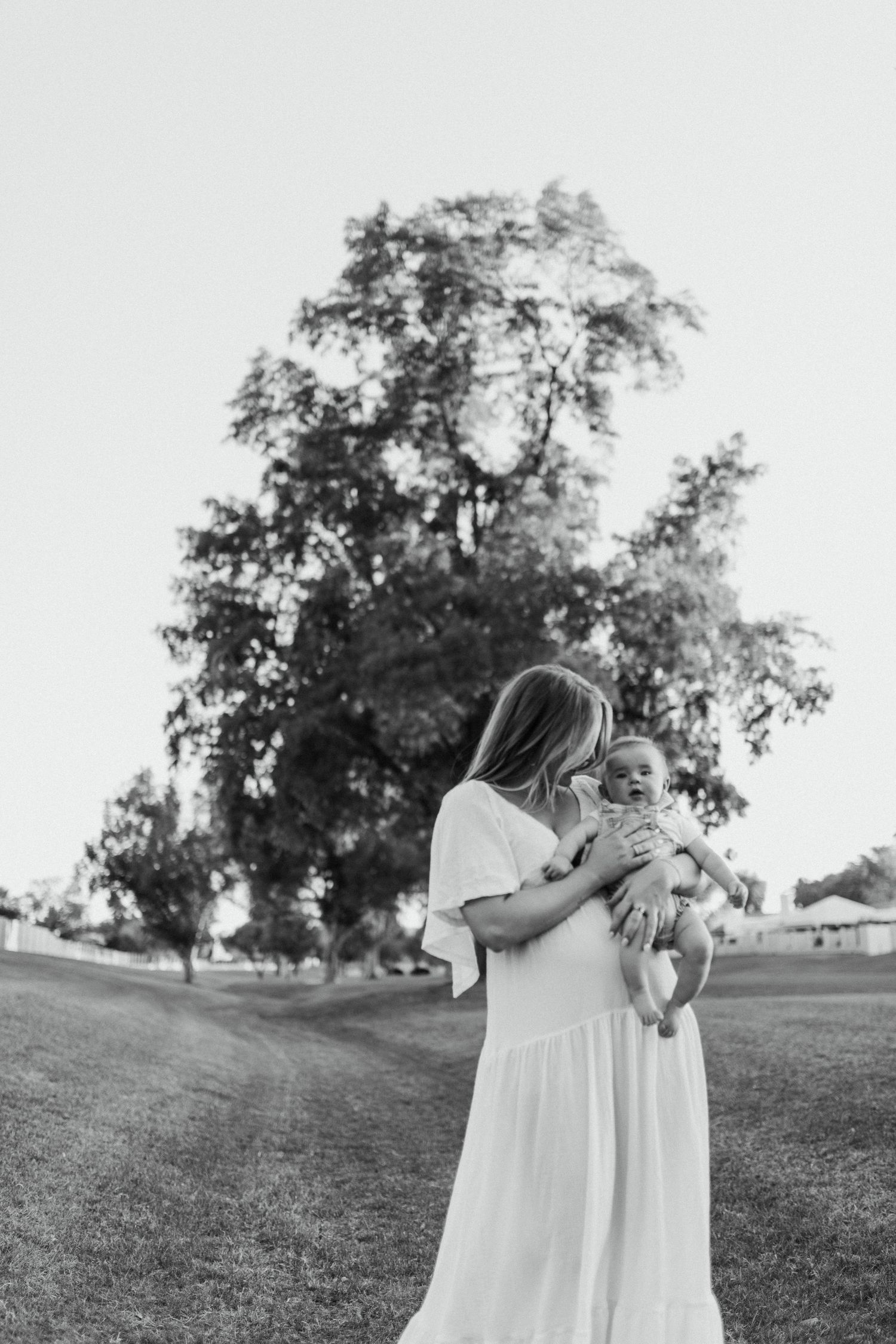 A tender moment captured in black and white as a parent in white dress holds a baby on a grassy lawn.