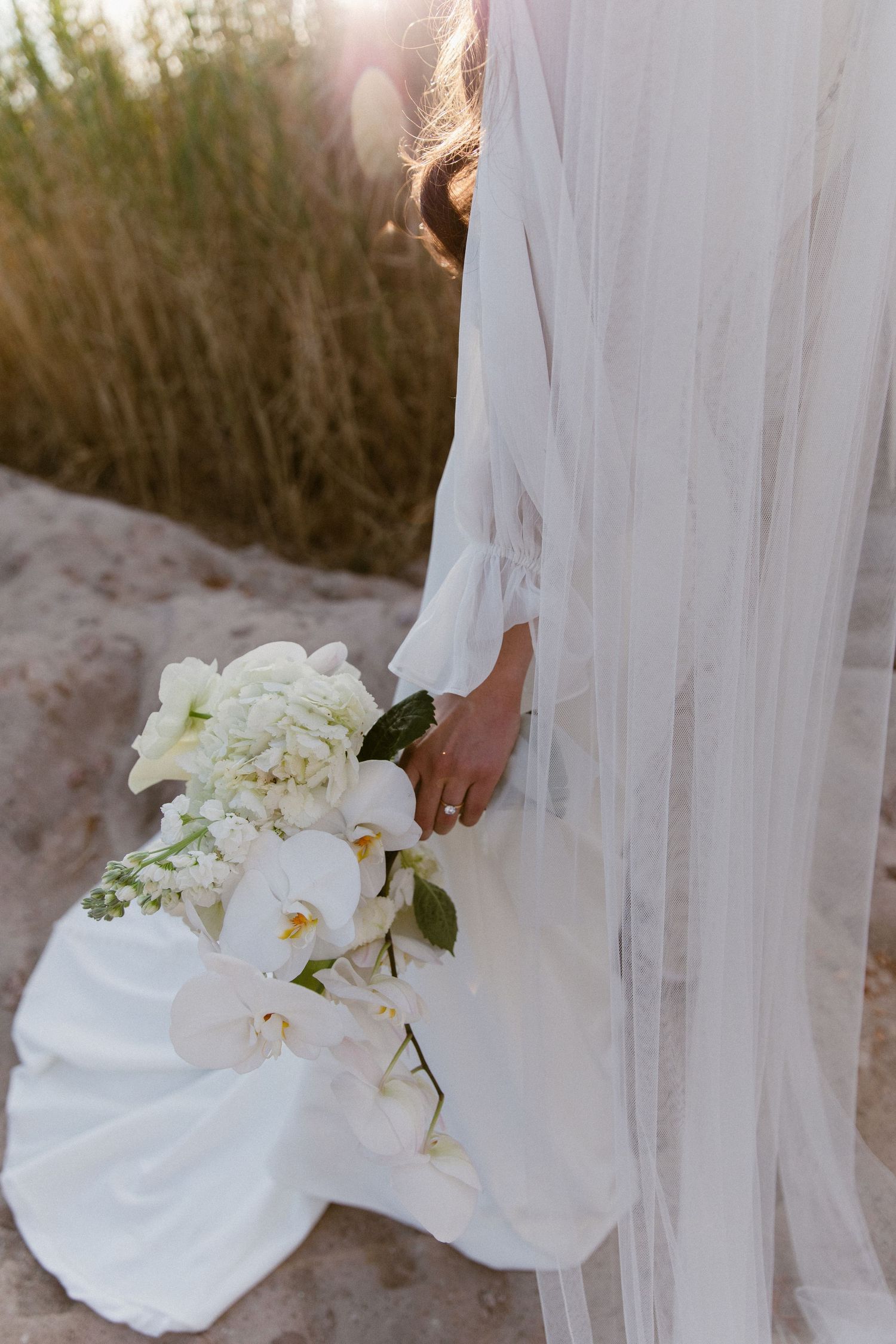 White bridal bouquet with orchids and roses resting against flowing wedding dress outdoors.