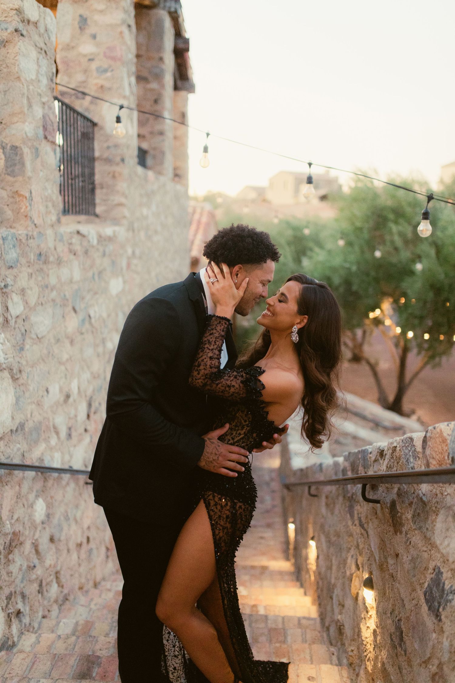 A romantic couple in elegant evening wear embraces on stone steps at dusk with string lights glowing in background.