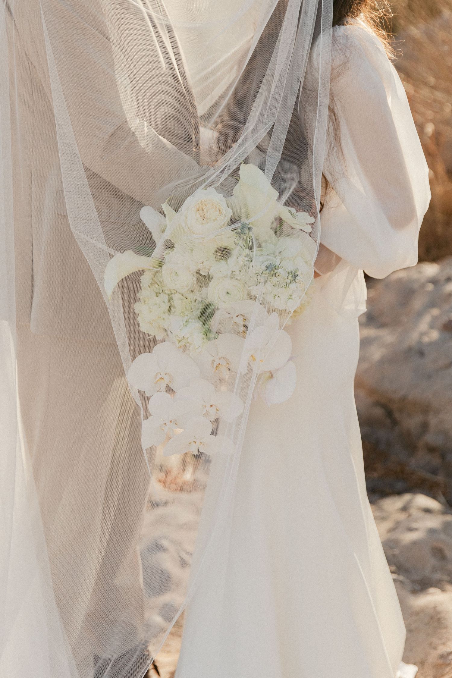 Bridesmaids in flowing white dresses hold delicate white floral bouquets during an outdoor wedding ceremony.