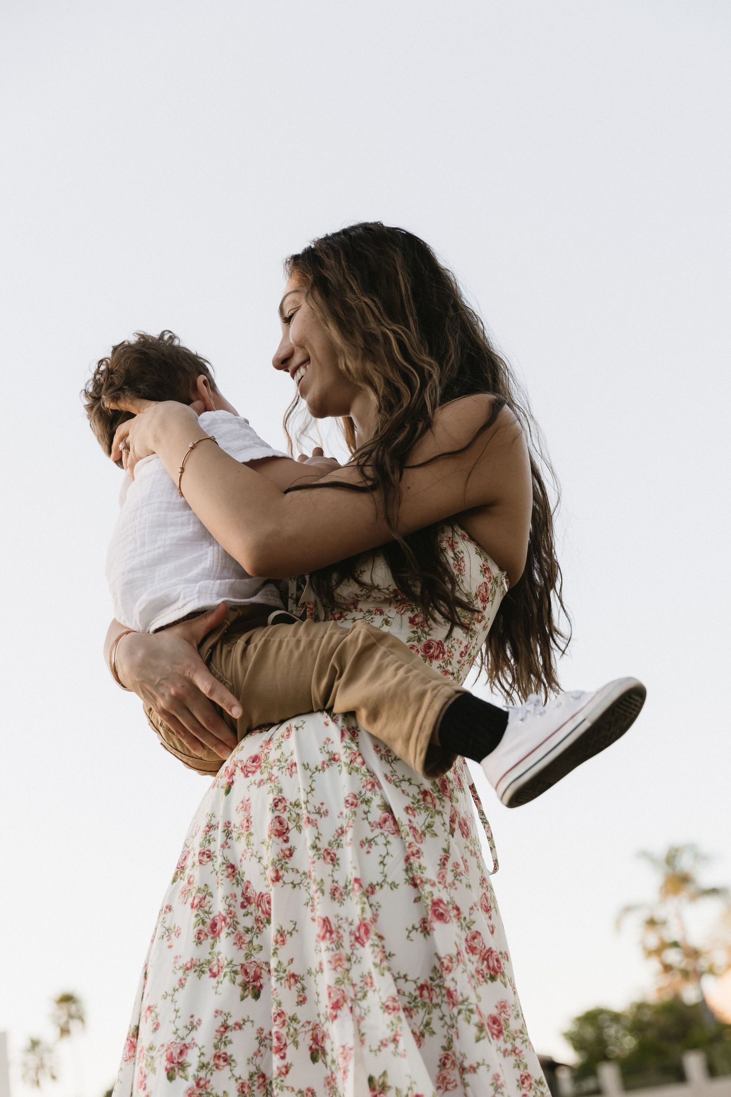 A woman in a floral dress joyfully lifts a child outdoors, both smiling and embracing against a soft sky.