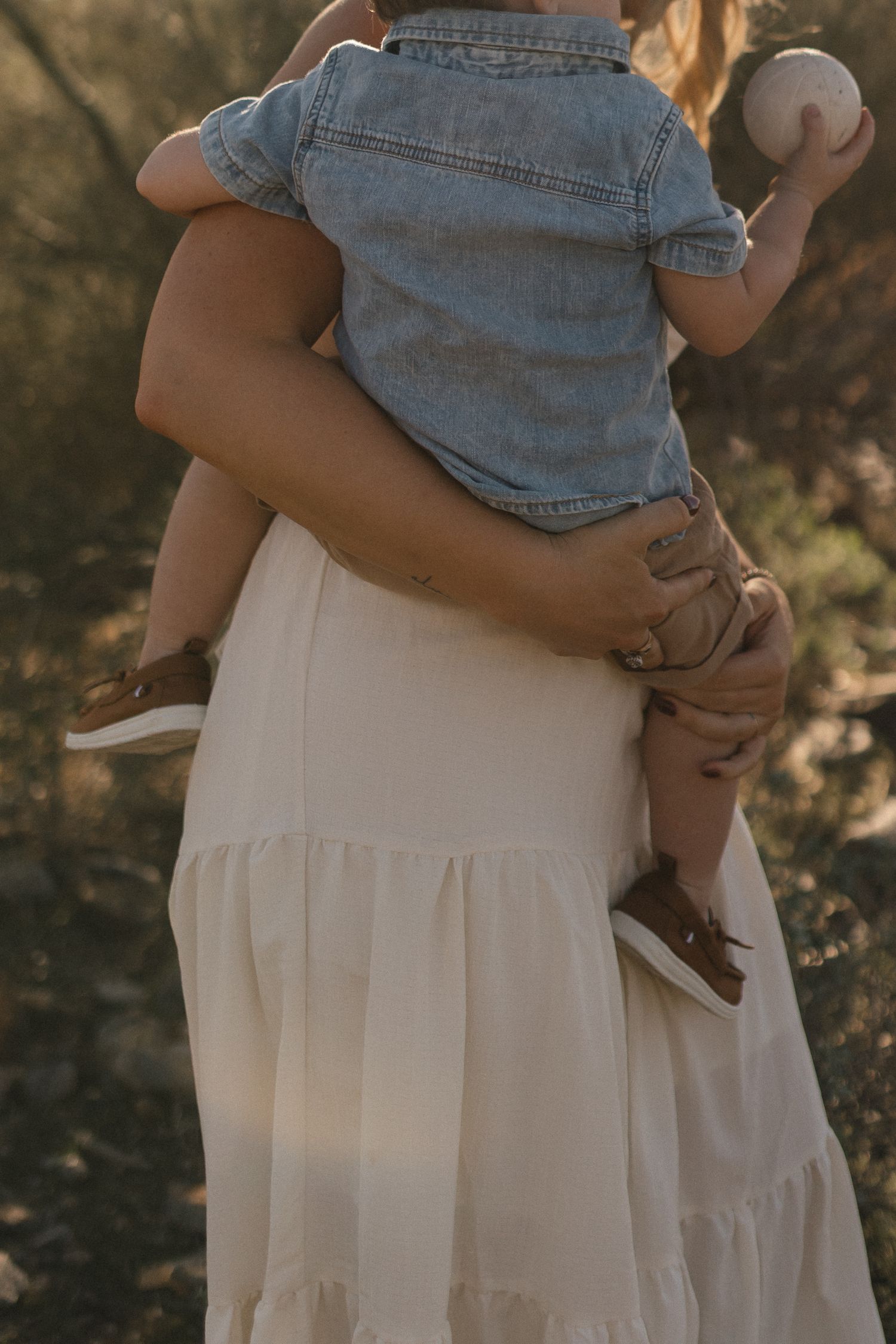A tender moment captured in warm light showing someone holding a baby wearing denim against a cream-colored dress.