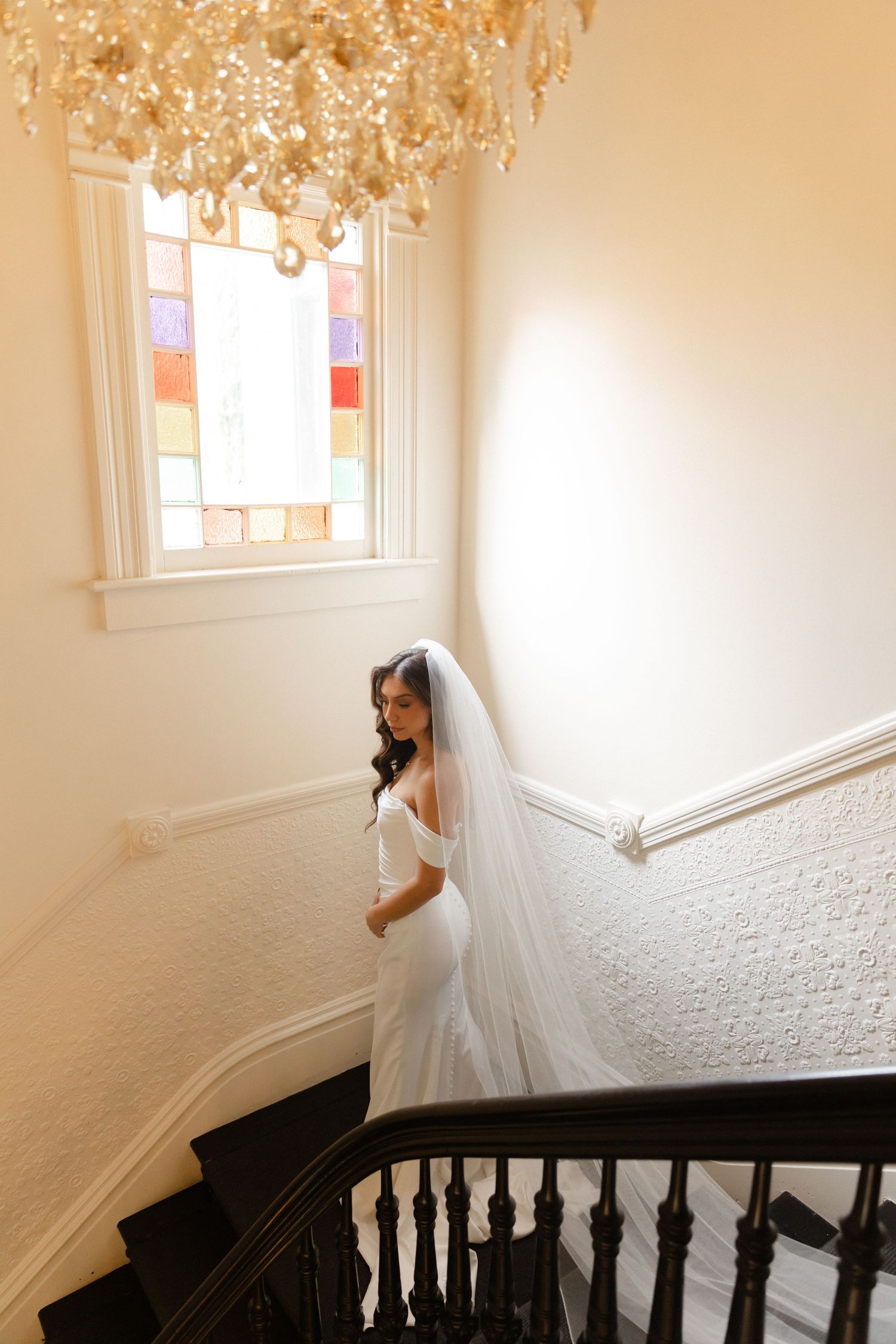 A bride in a flowing white wedding dress descends an elegant curved staircase with dark wooden railings.