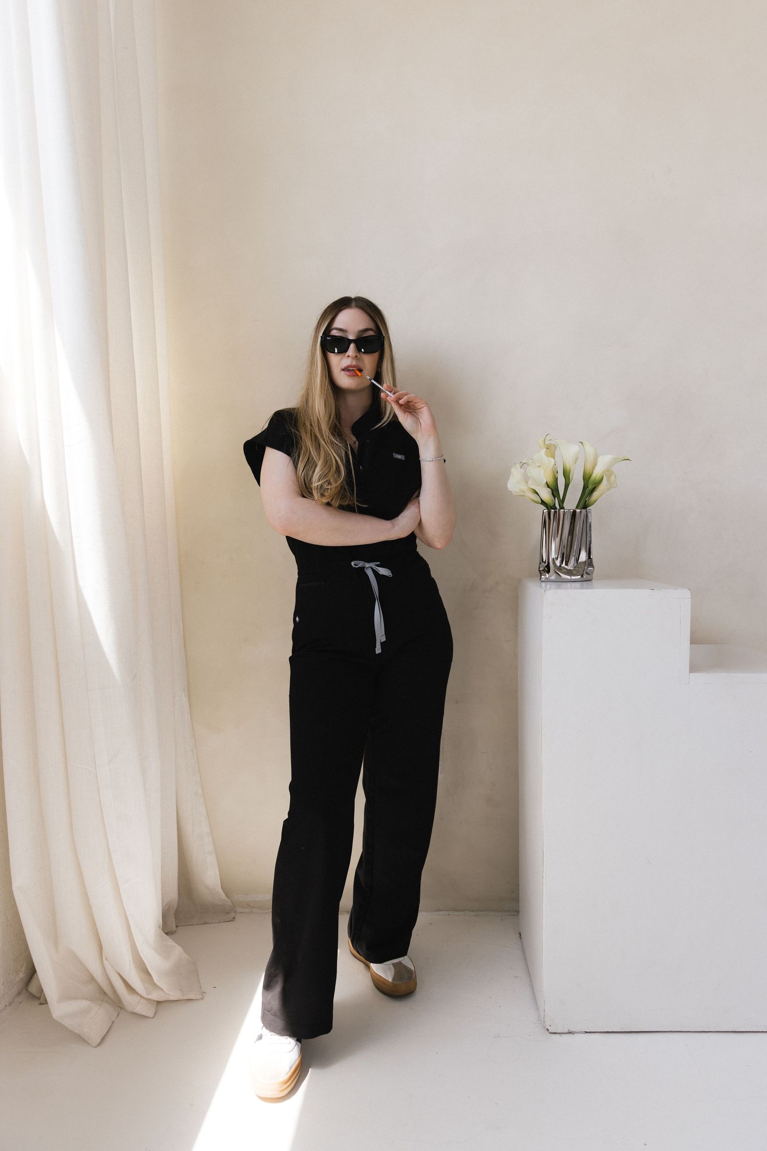 Woman in black outfit and sunglasses poses near white pedestal with flowers in bright minimalist room.