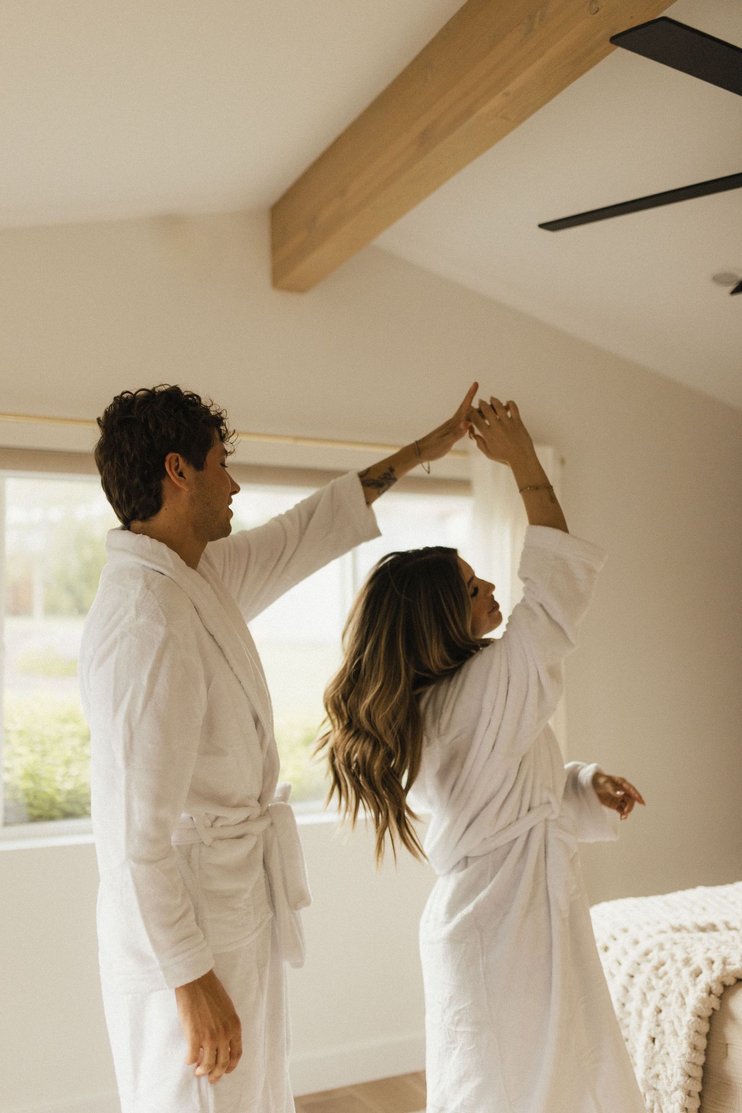 Couple in white robes dancing together in a bright bedroom with wooden ceiling beams.