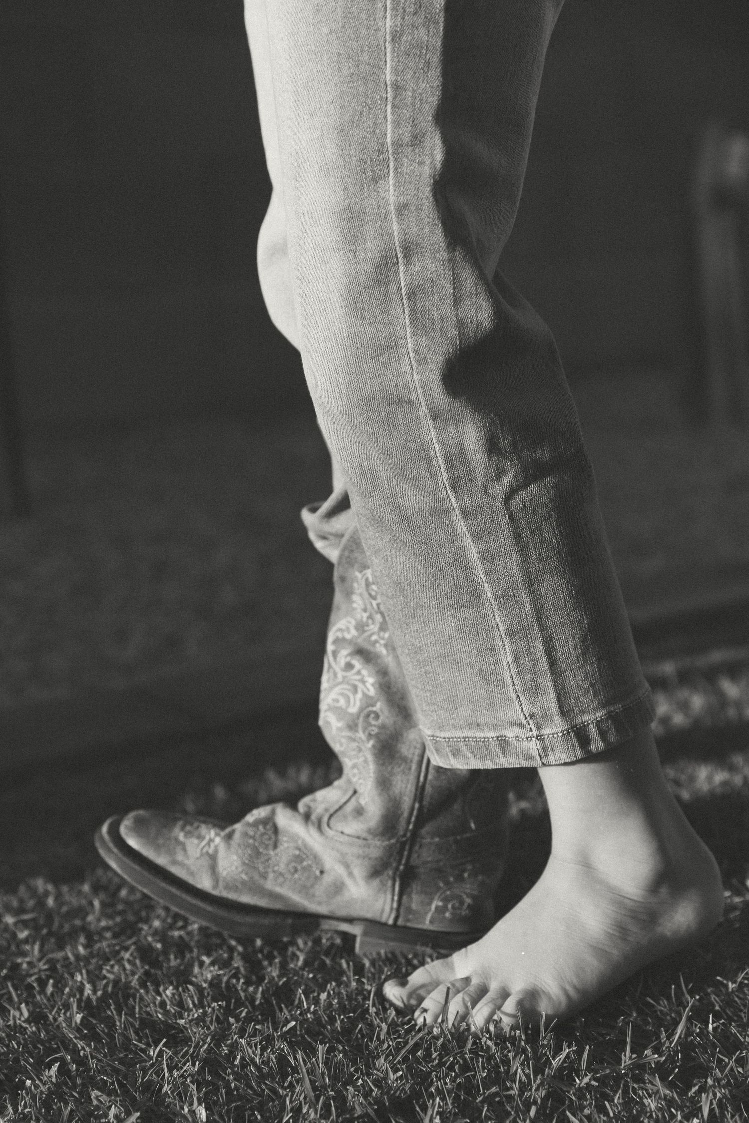 Black and white photo of person wearing cowboy boots and jeans standing barefoot on grass.