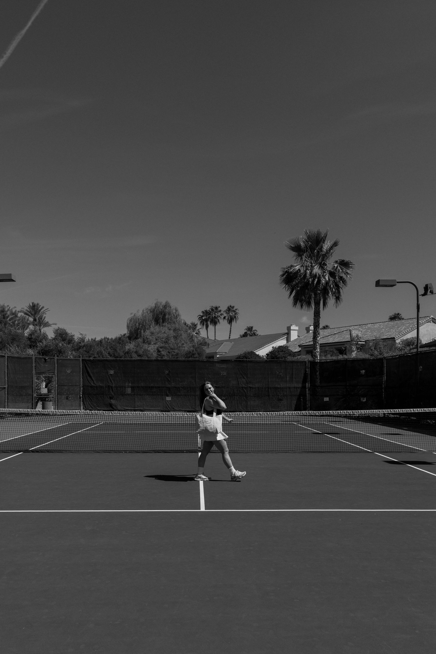 Black and white photo of a tennis player mid-swing on an outdoor court with palm trees in the background.