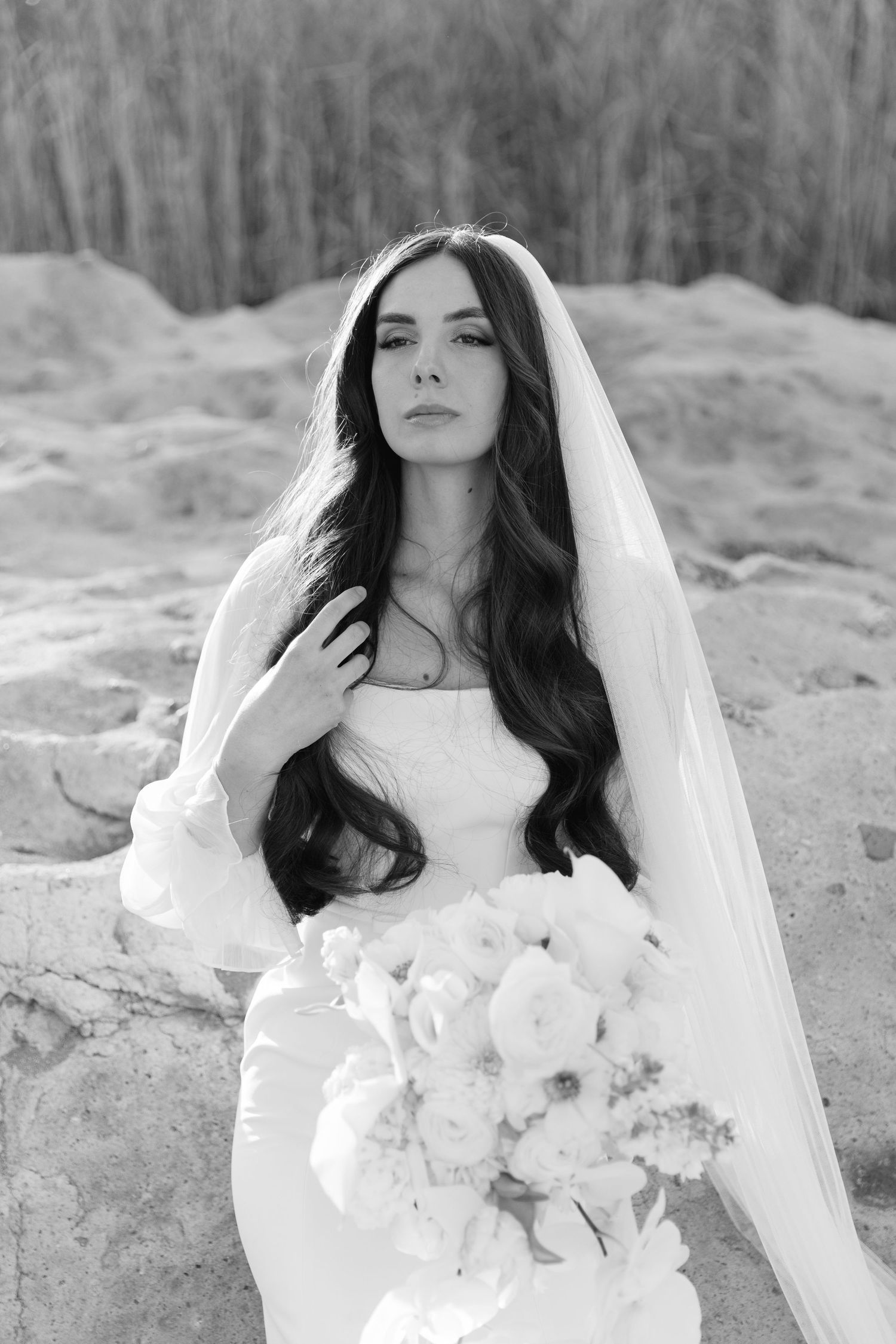 A bride in a white wedding dress holds a bouquet of white flowers against a rocky outdoor backdrop.