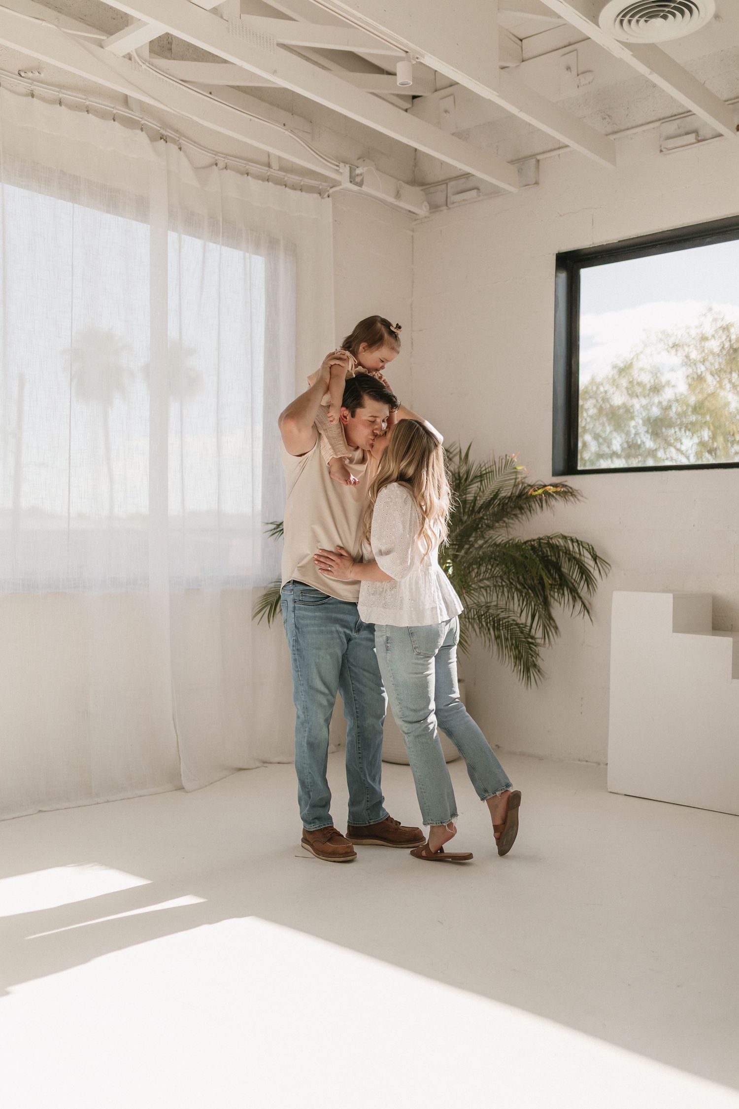 A couple embraces in a minimalist white studio space with sunlight streaming through sheer curtains.