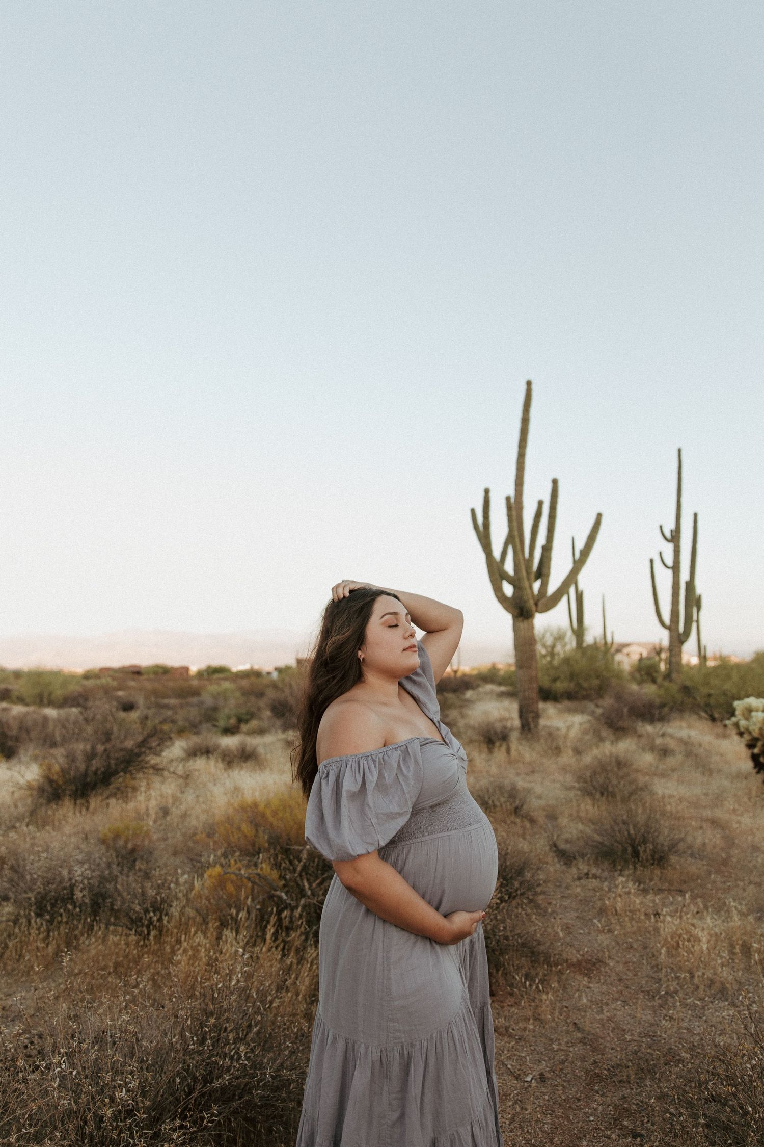 A maternity photo of a woman in a flowing gray dress posing against towering saguaro cacti.