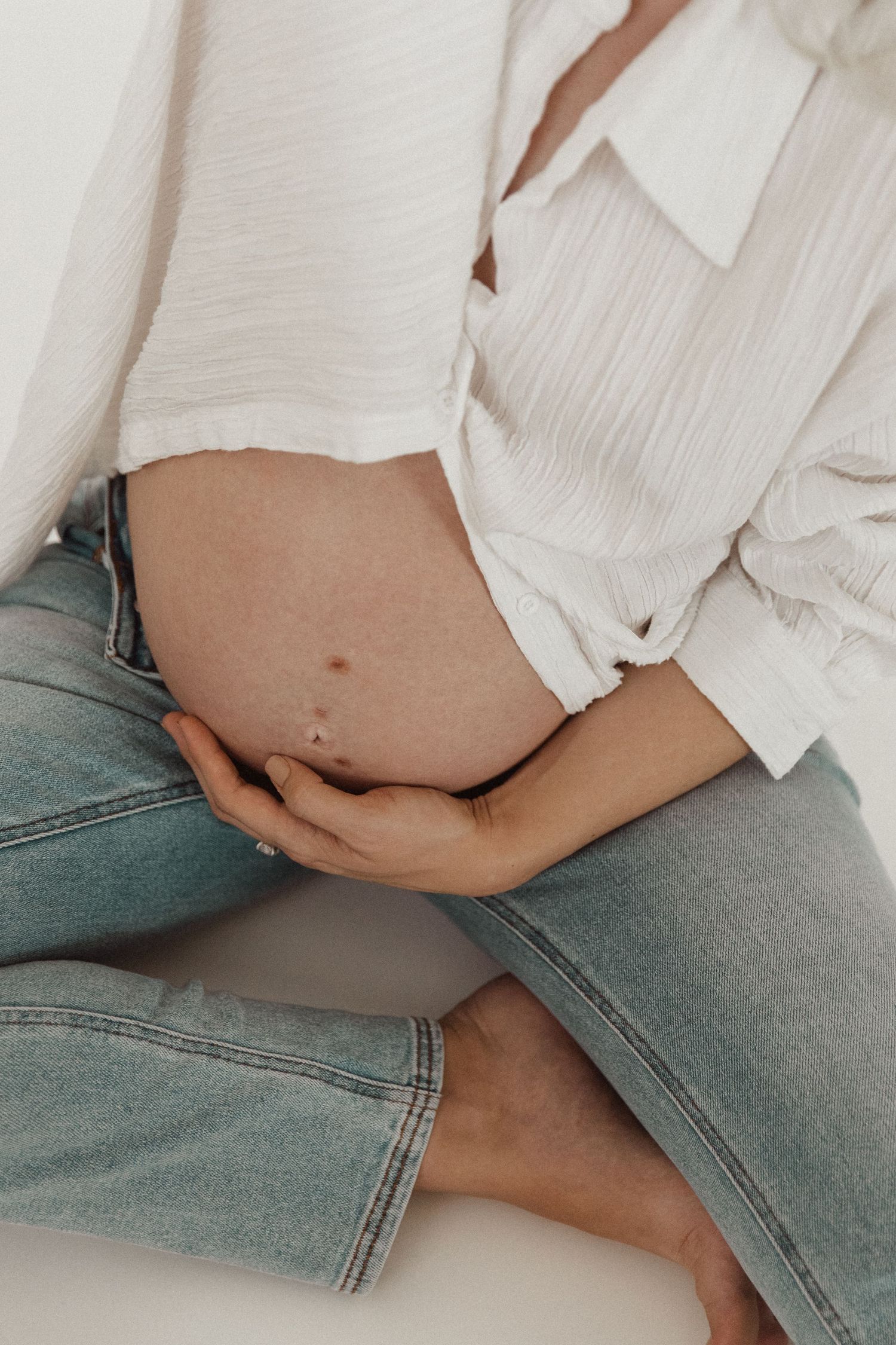 Close-up of a pregnant belly with hands cradling it, wearing jeans and white shirt.