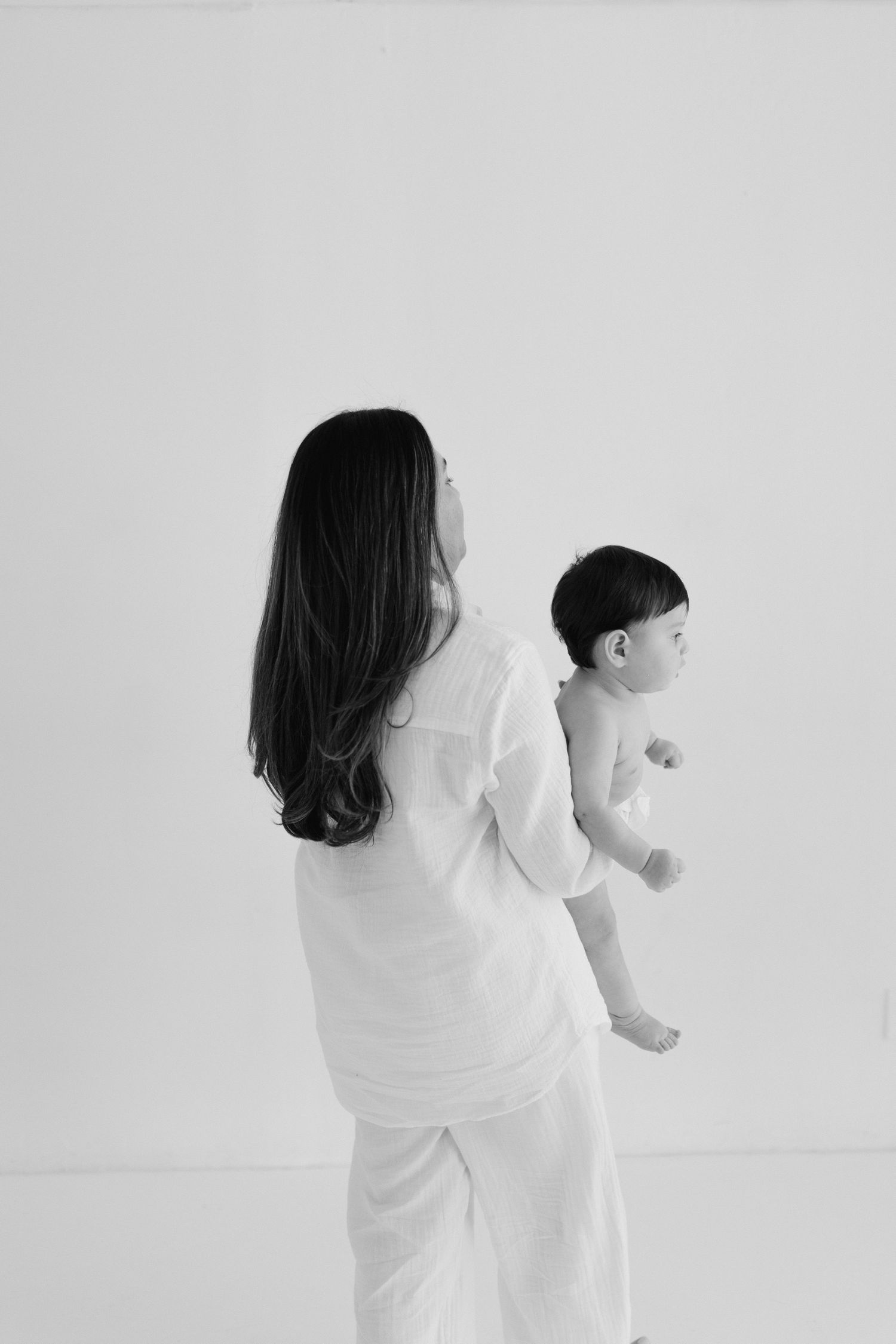 Black and white photo of a mother in white clothing holding her baby against a light background.