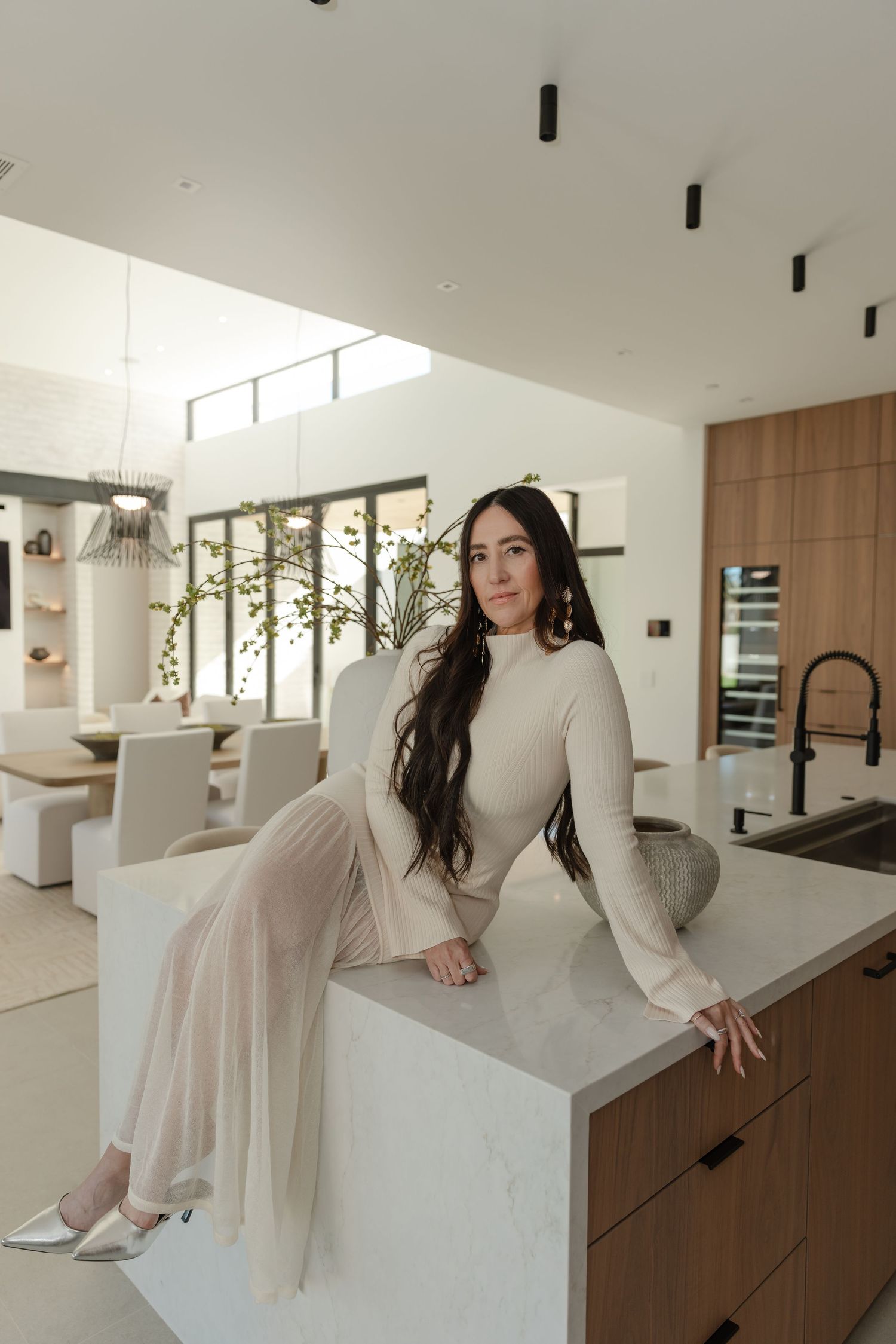 Woman in beige turtleneck dress sits on marble kitchen island in modern luxury home with wood accents.