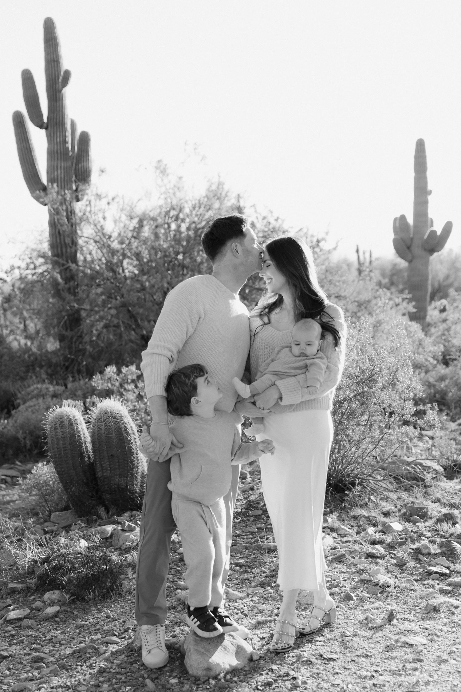 A family shares a tender moment in a desert landscape with towering saguaro cacti in black and white photography.