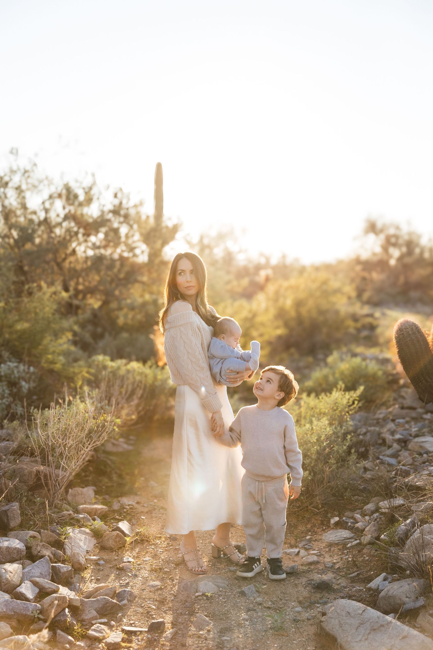 Two figures stand together in a desert landscape at golden hour with sunlight streaming through cacti and rocks.