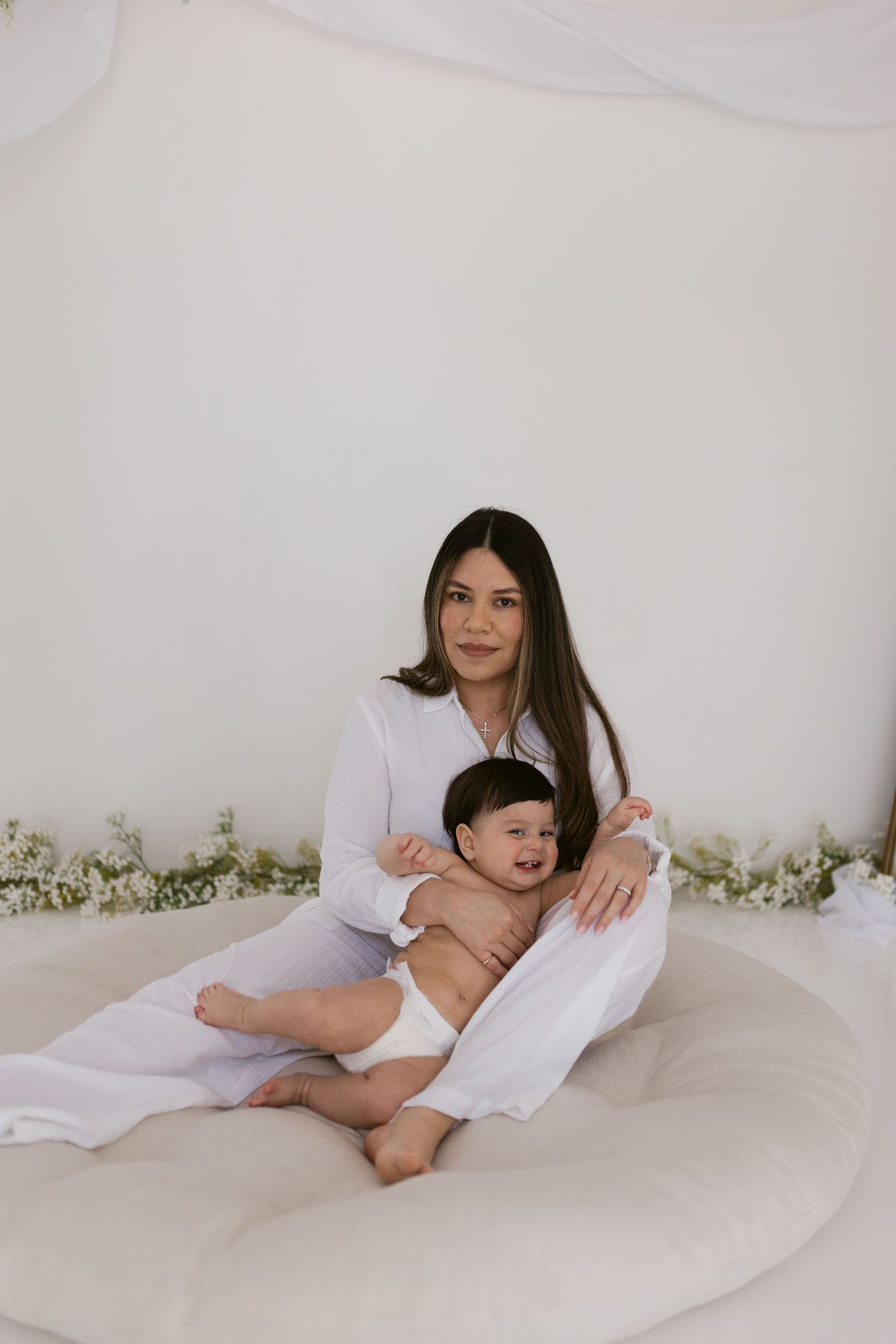 A young mother in white sits on a round cushion holding her smiling baby, surrounded by white flowers and draping.