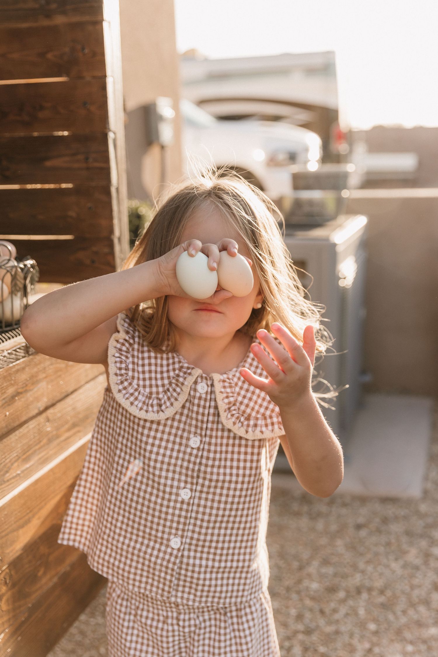 Young girl in pink gingham dress holding two white eggs over her eyes outdoors in warm golden sunlight.