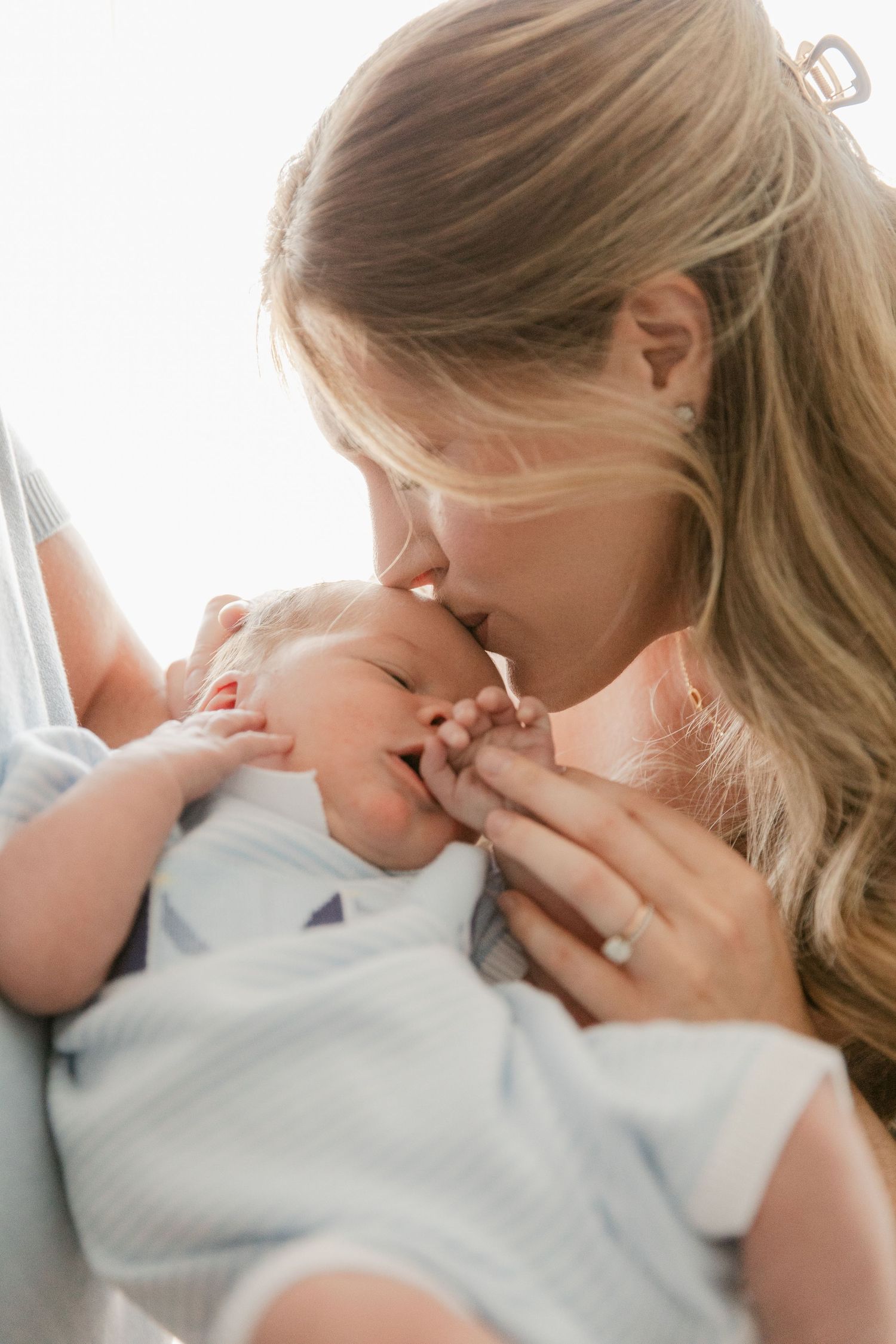 A tender moment between a mother and sleeping newborn baby as she gently kisses the infant wrapped in white.