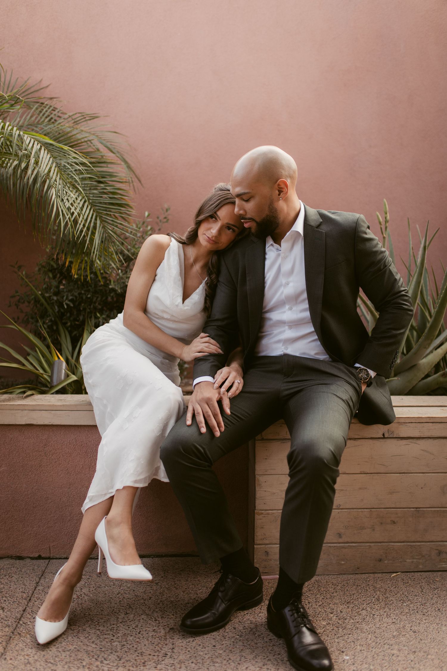 A couple in wedding attire shares an intimate moment on a bench against a terracotta wall with palm fronds.