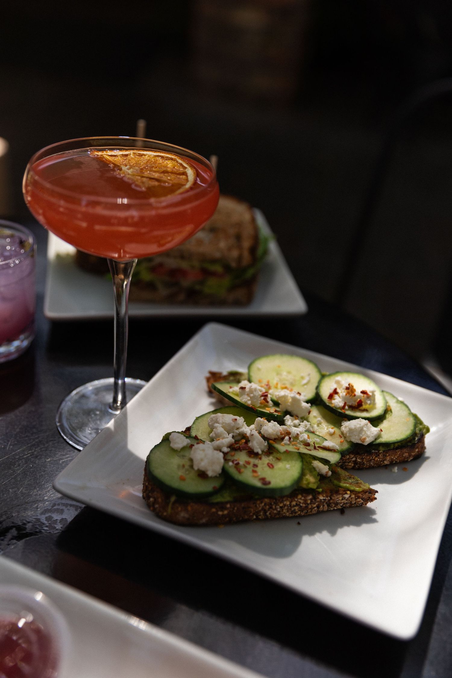 Red cocktail and open-faced cucumber sandwiches on dark bread served on white plate.