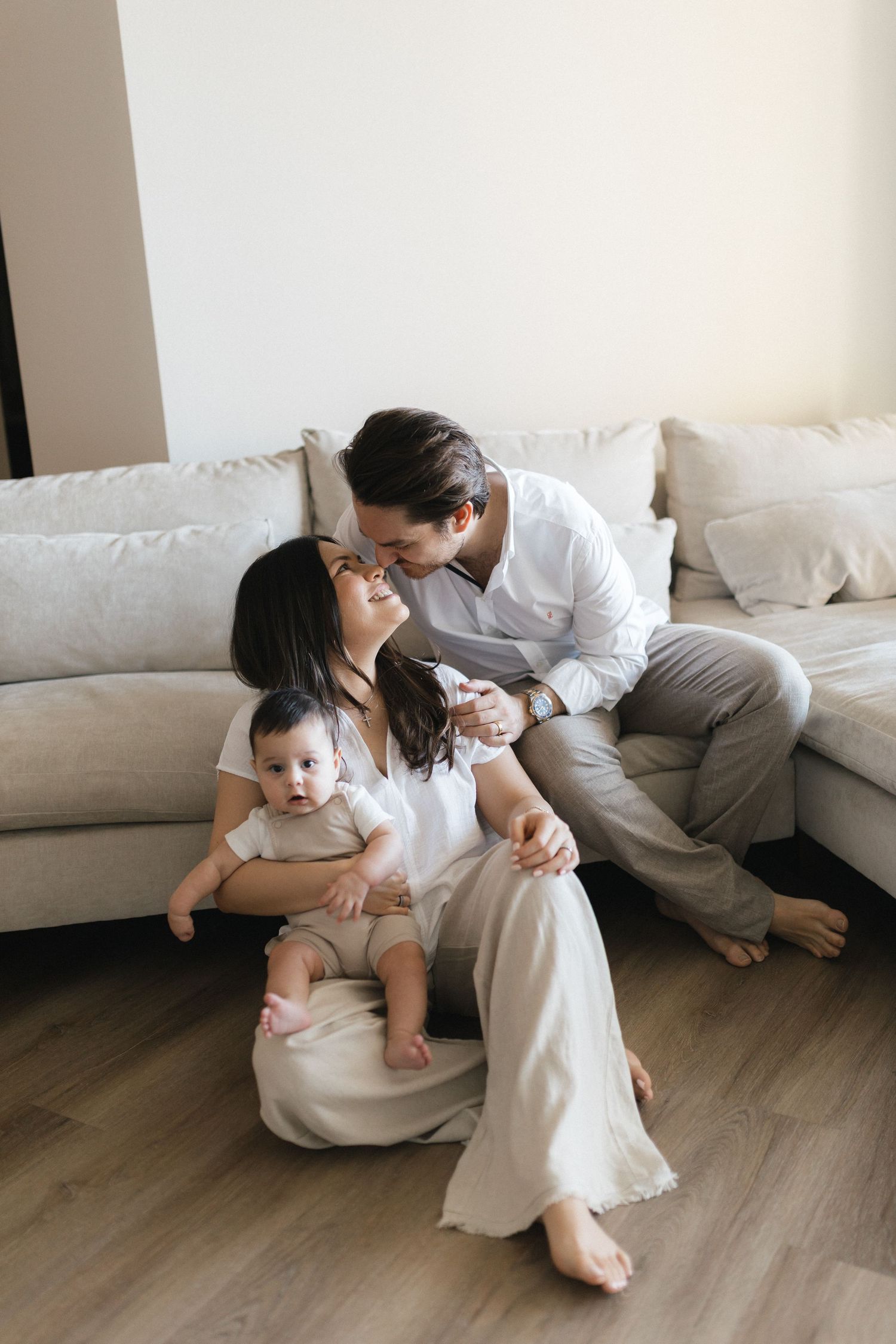 Family relaxing together on light hardwood floor beside white couch in minimalist home setting.