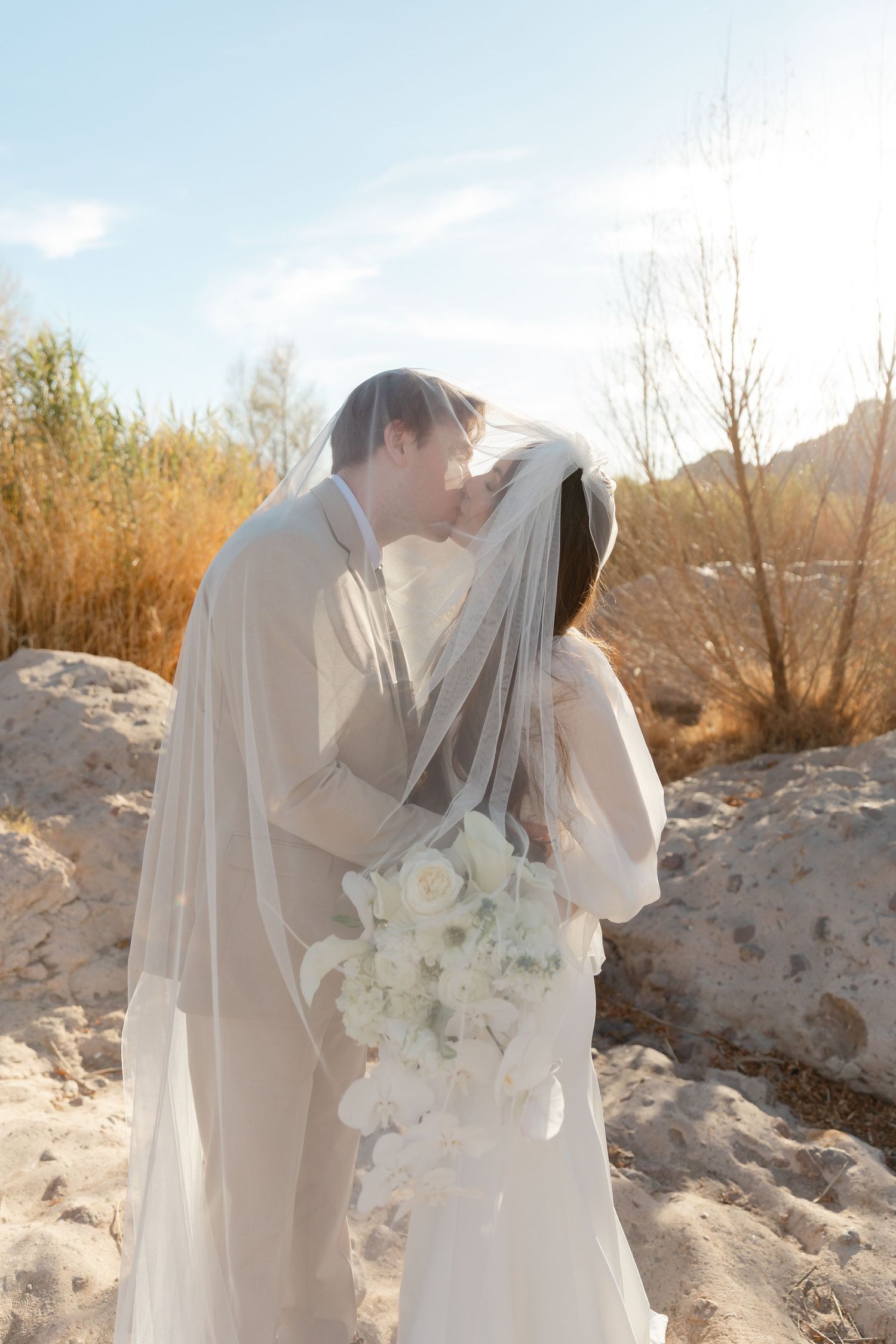 Romantic wedding couple embracing outdoors in a desert setting with white floral bouquet.