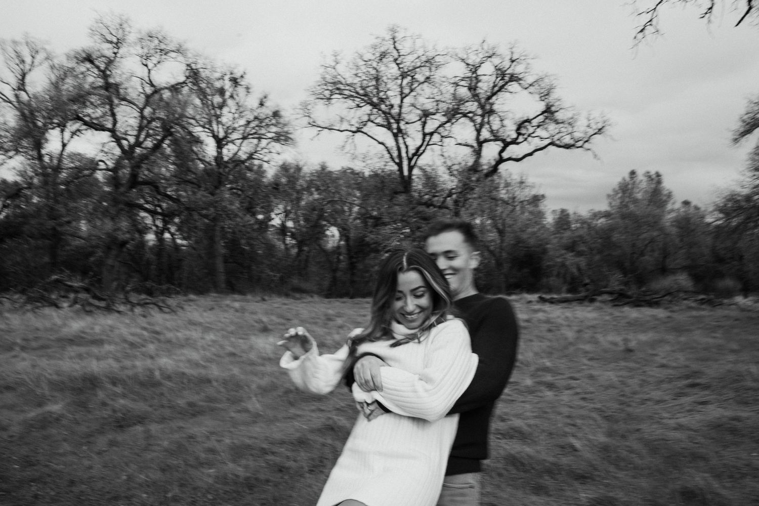 A romantic black and white photo of a couple embracing in a field with trees.