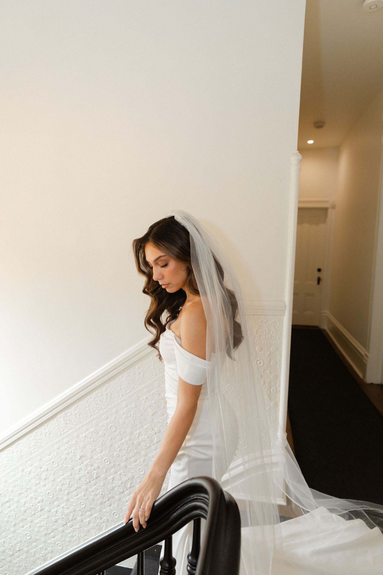 A bride in a white off-shoulder wedding dress and long veil stands elegantly by a staircase railing.