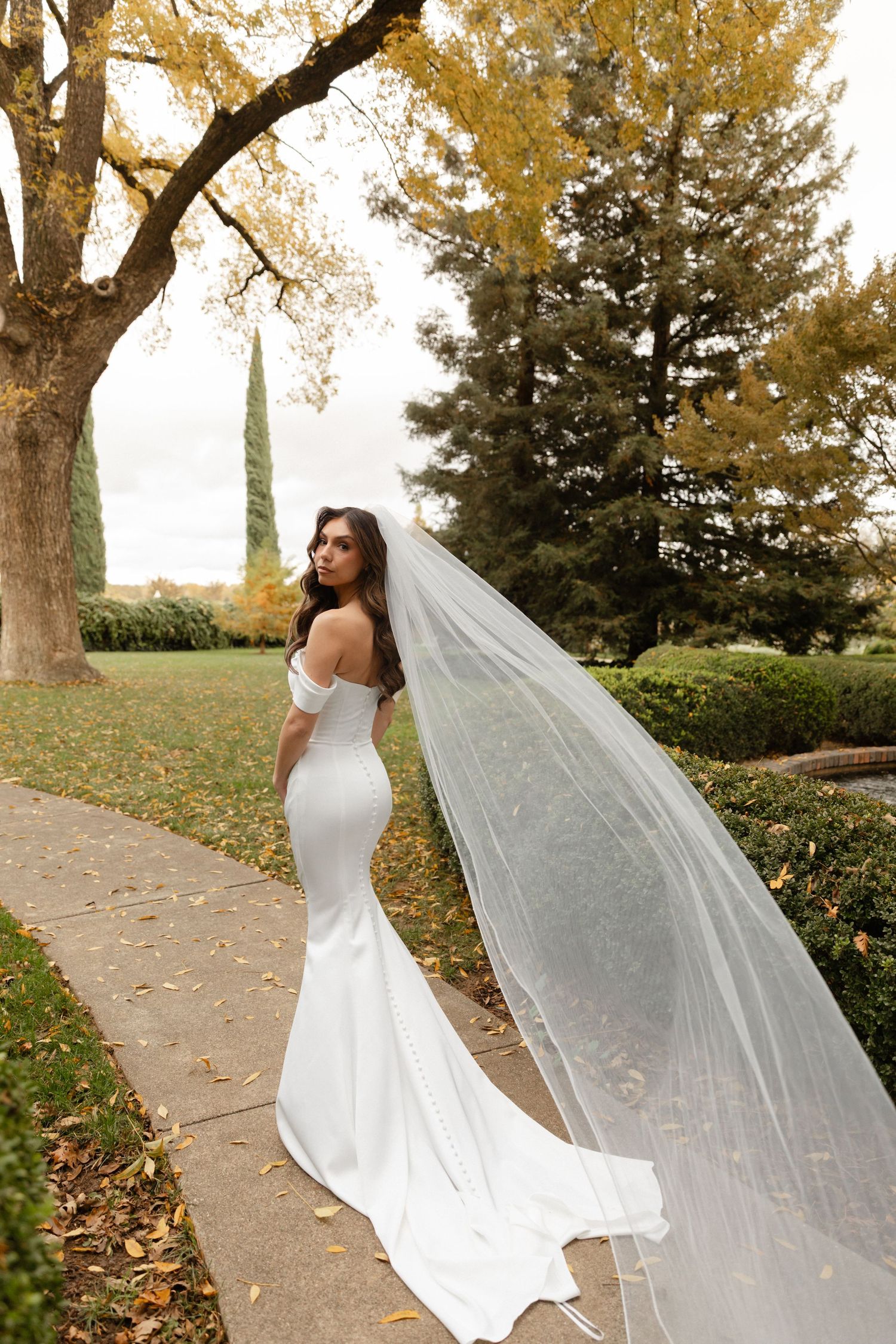 A bride in a white wedding dress poses with a flowing veil in a scenic autumn park setting.