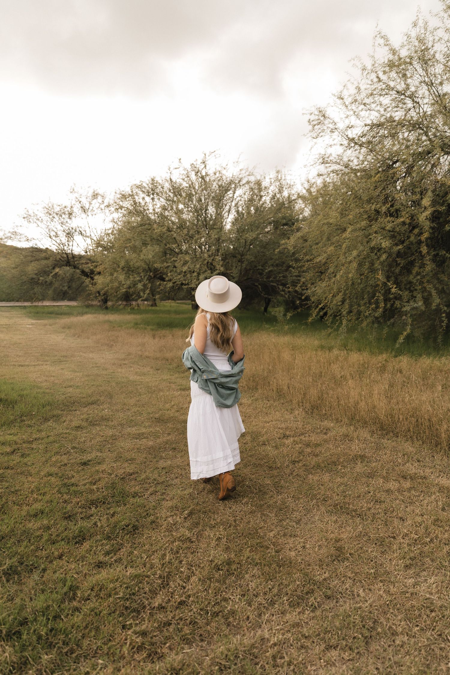 A person in a white dress and sun hat stands in a grassy field with trees in the background during golden hour.