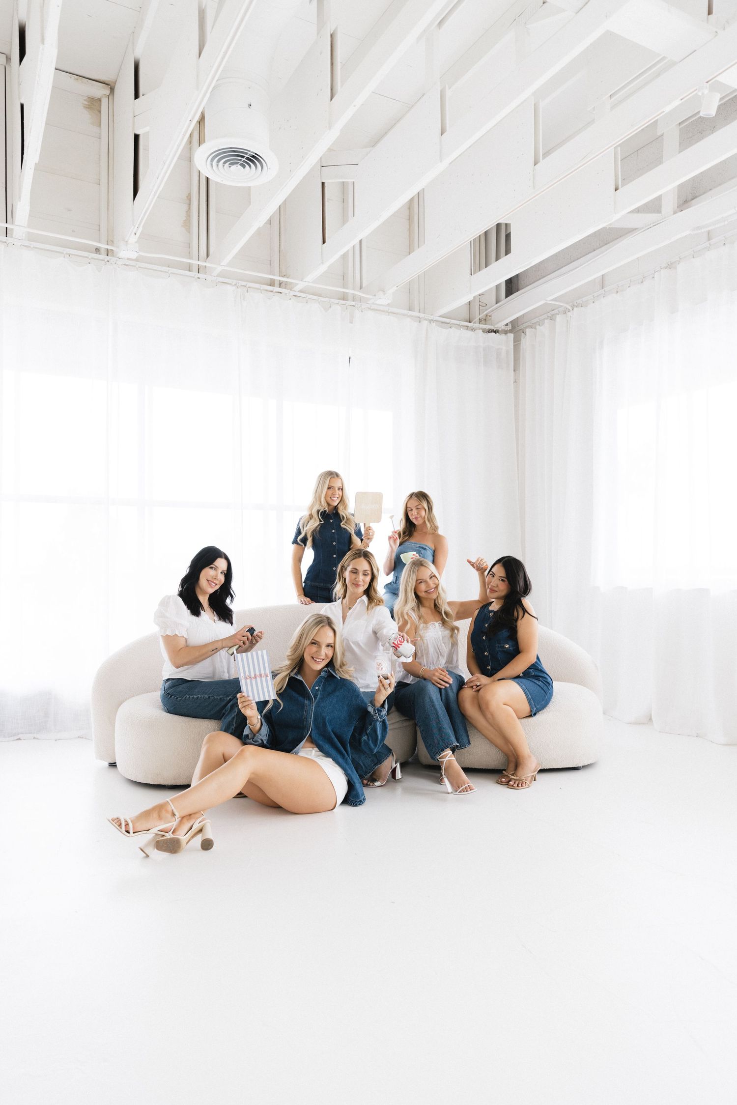 Seven women in denim and white outfits pose together in a bright white studio space for a stylish group photo.