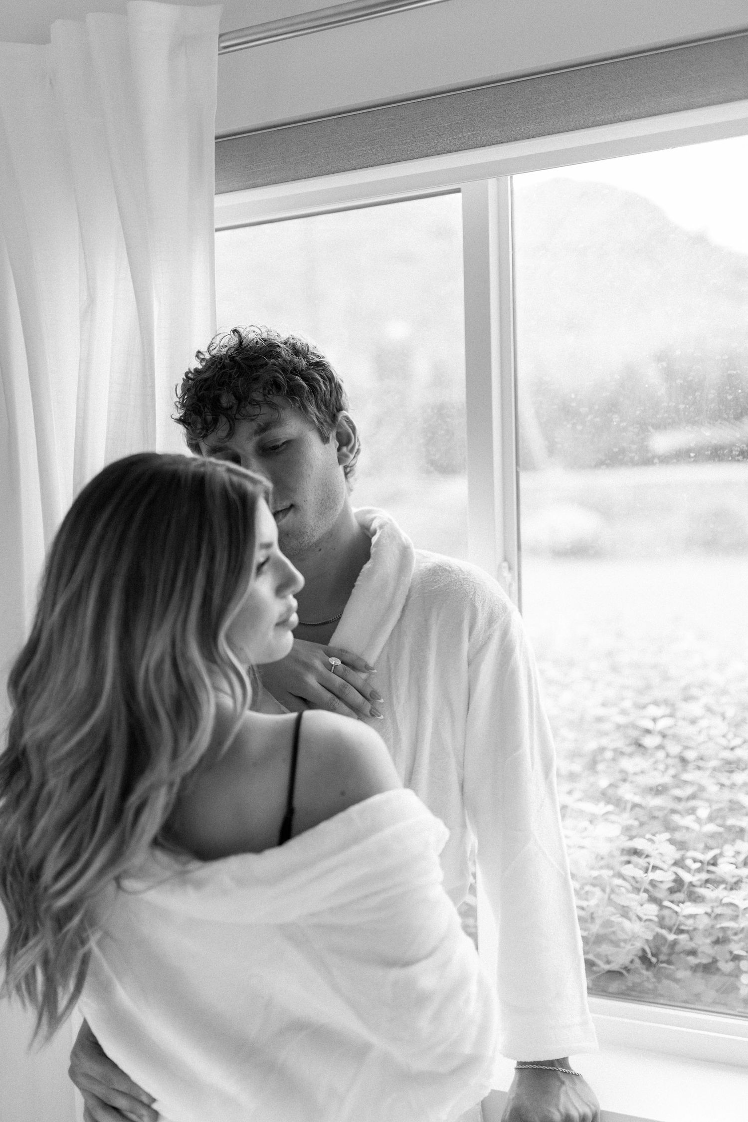 Black and white photo of a romantic couple in white robes embracing by a large window with soft natural light.