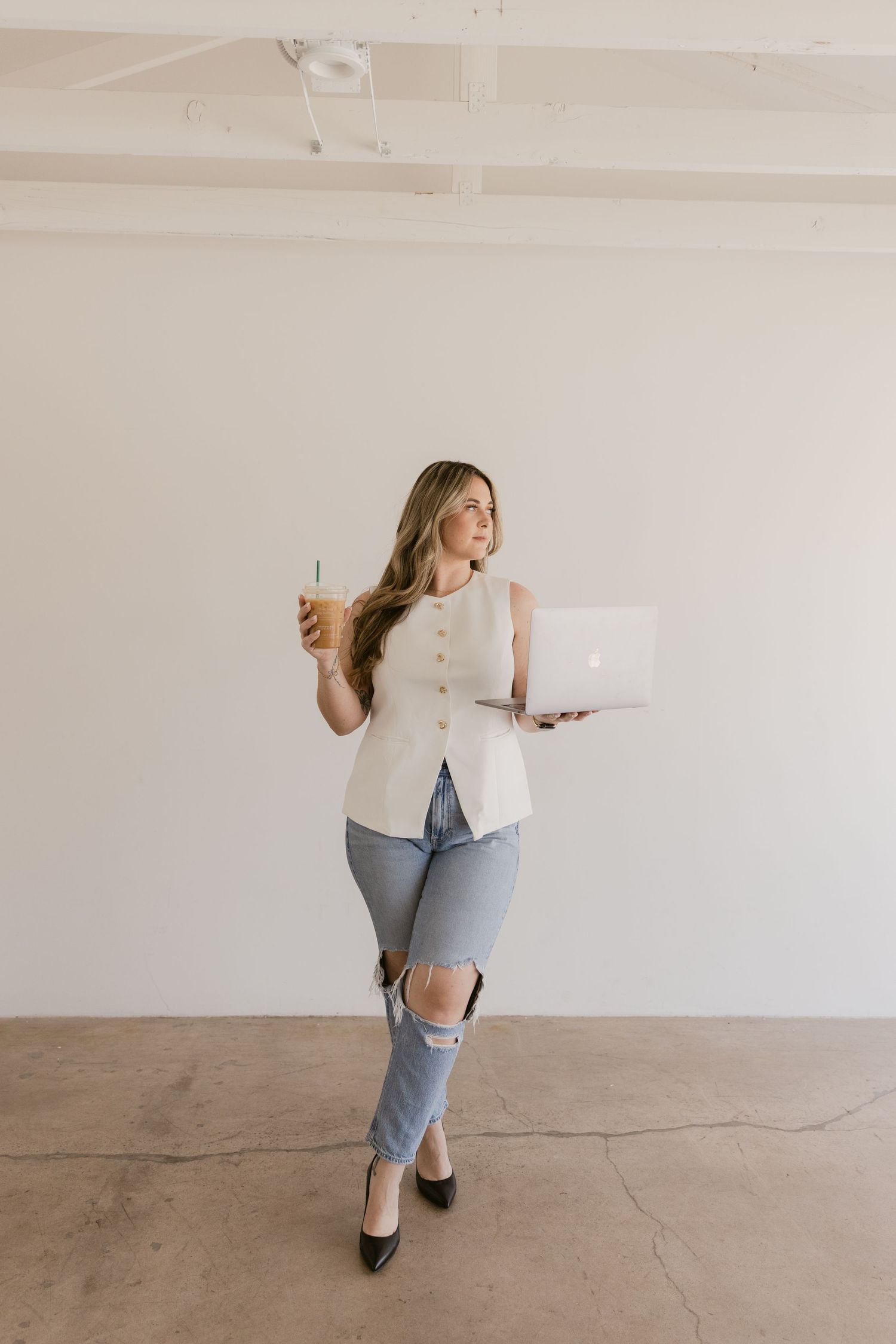 Woman in white sleeveless top and ripped jeans holding laptop and iced coffee, standing in minimalist white studio space.