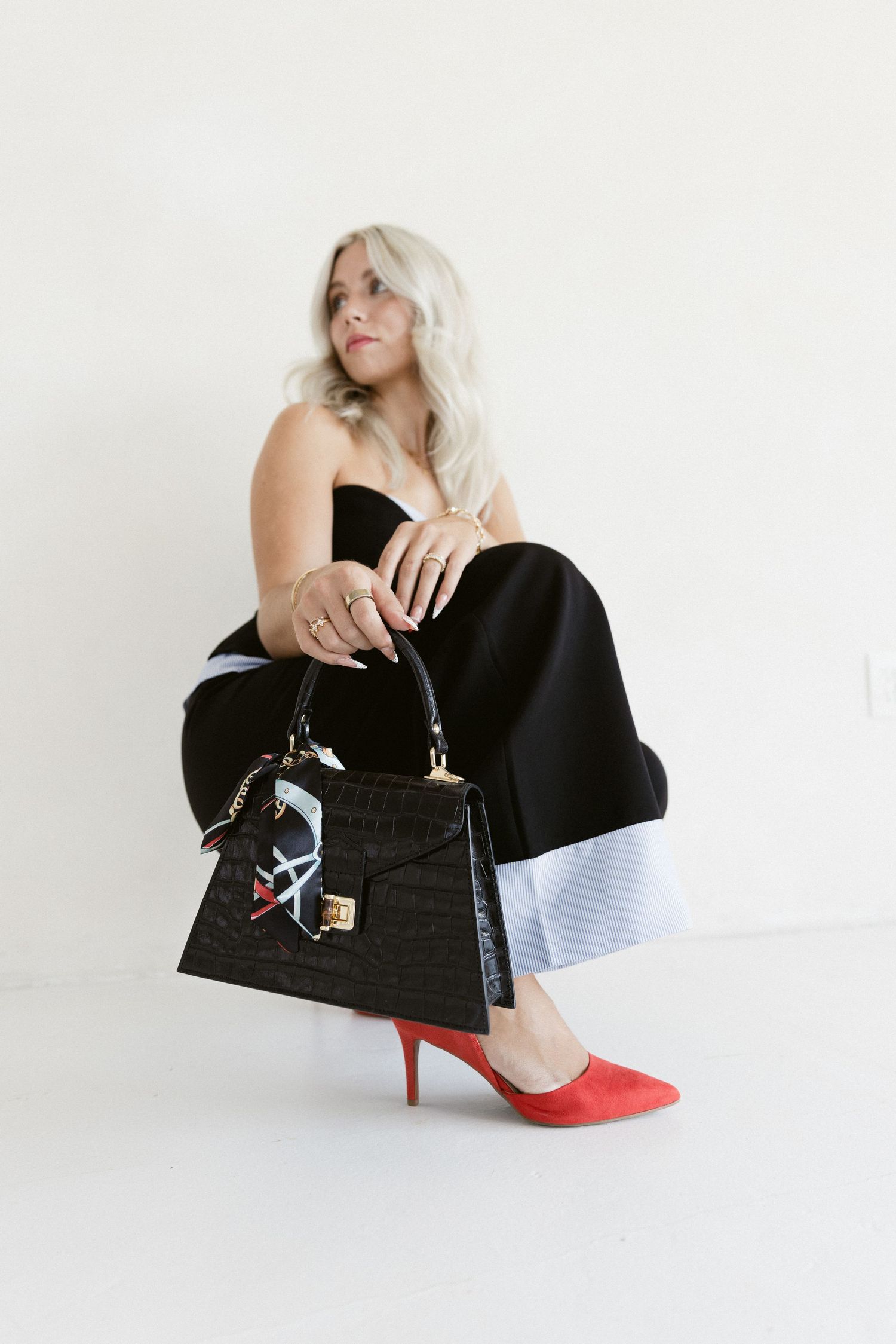 Woman in black strapless jumpsuit with red heels holding a black croc-embossed handbag with scarf detail.