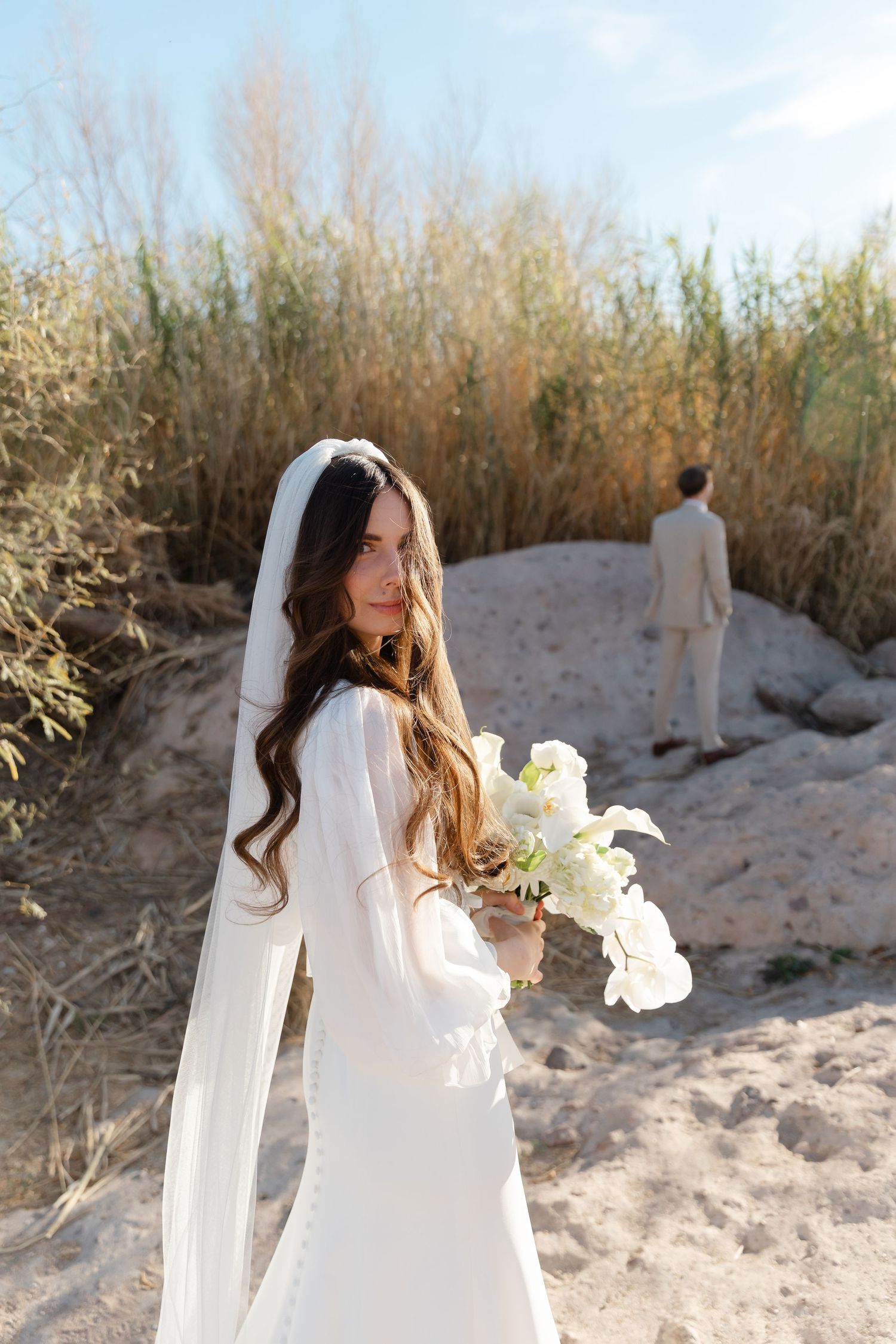 A person in a flowing white dress holds white flowers while standing on sandy terrain against tall dried grass.