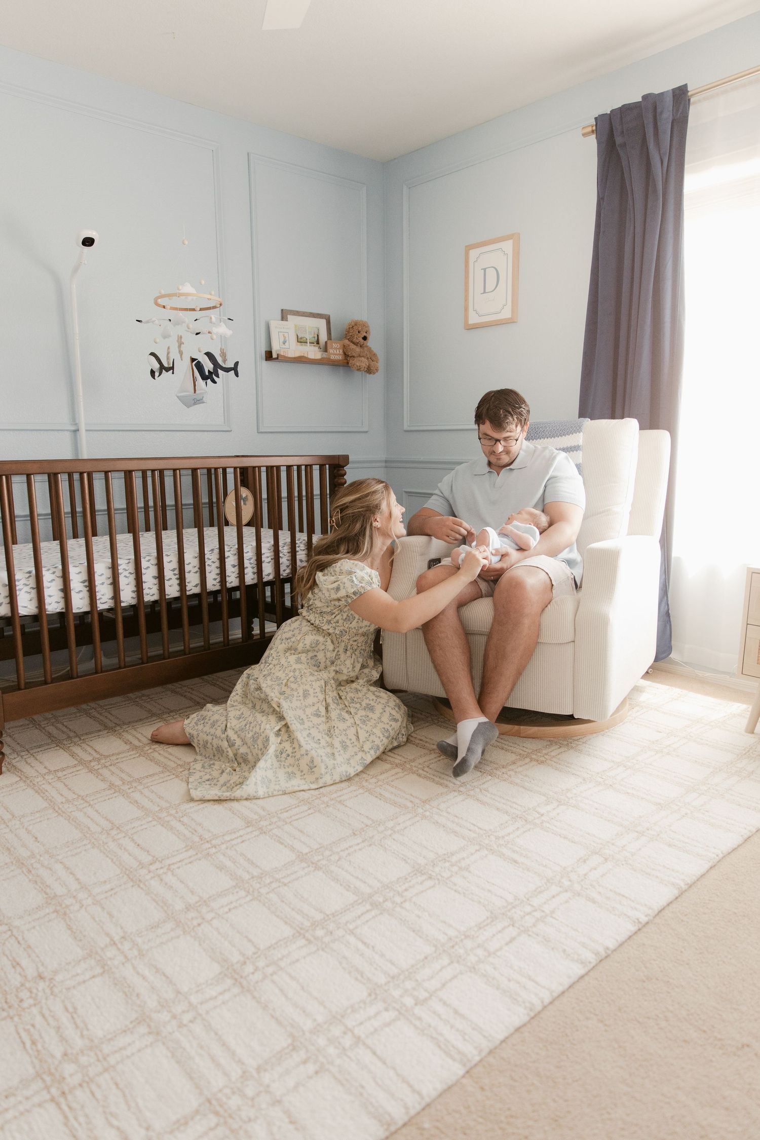 A nursery room with light blue walls features a wooden crib and a comfortable rocking chair where two people share a tender moment.