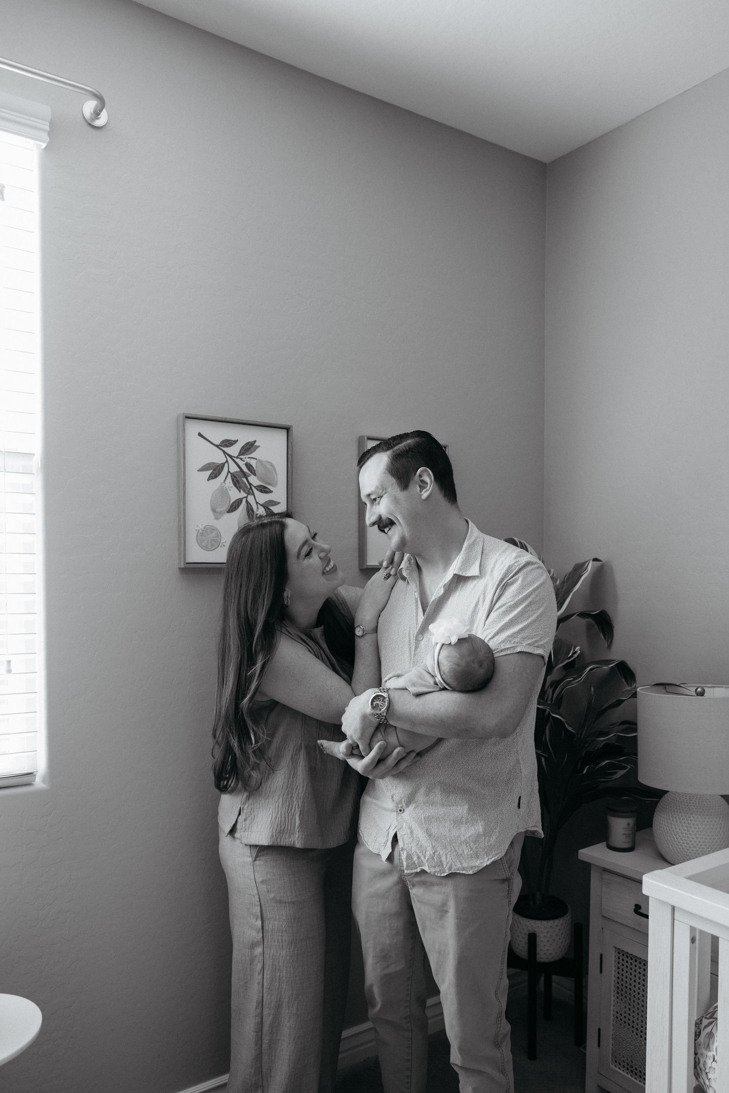Black and white photo of a couple smiling at each other while holding their newborn baby in a nursery room.