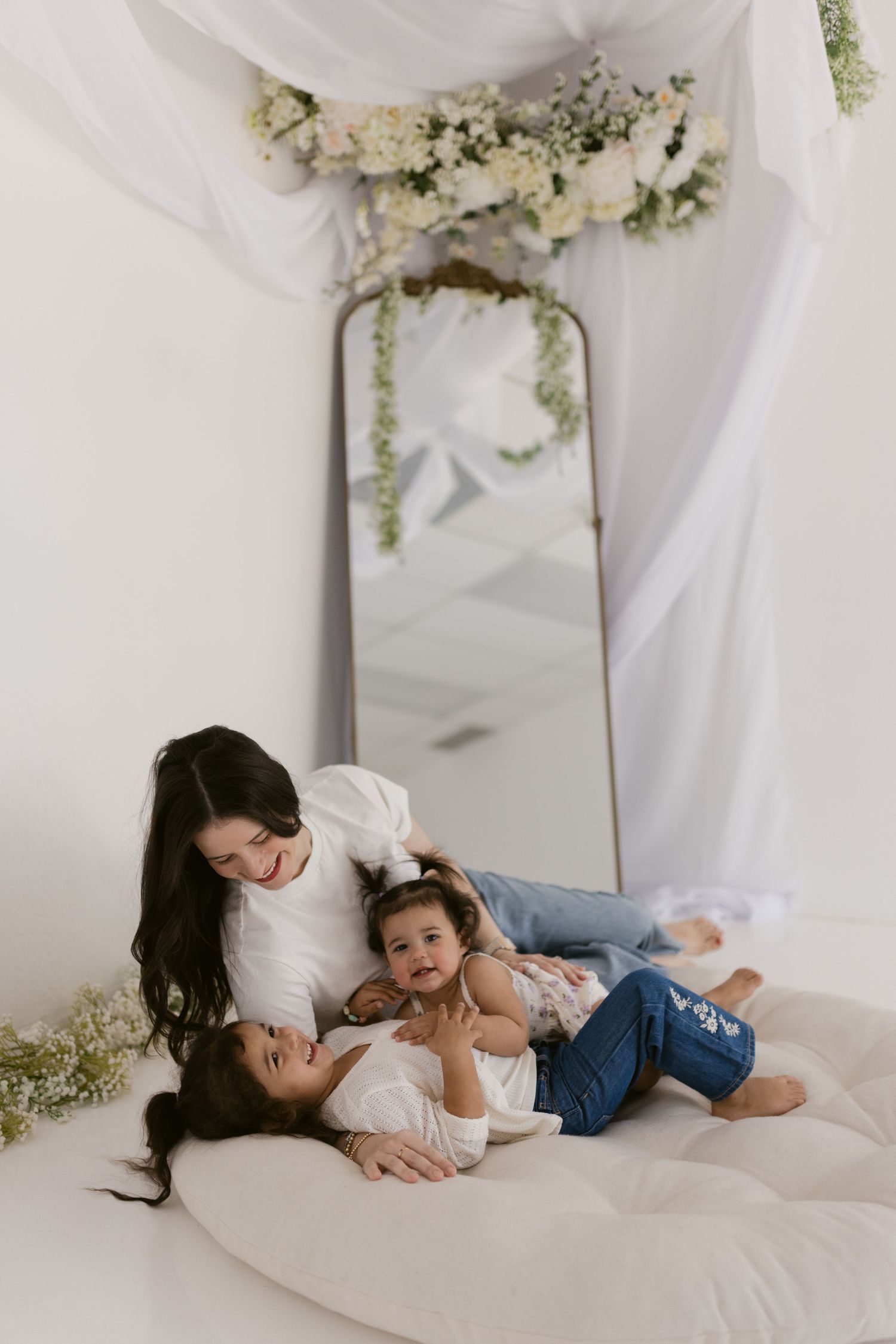A mother smiles warmly at her two young children while relaxing on a white cushion in an elegant floral studio setting.