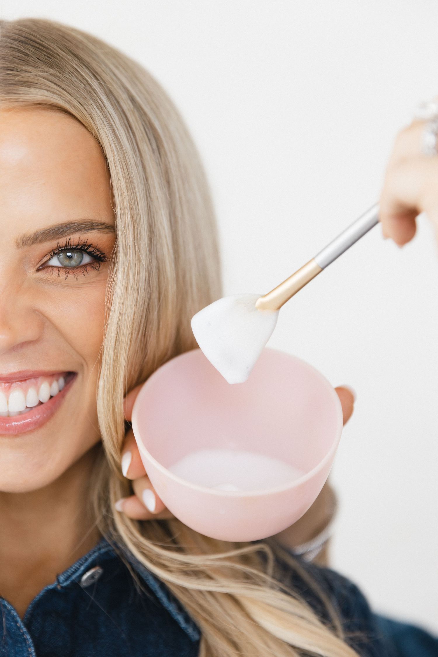 Smiling blonde woman holding a pink bowl and brush, applying a facial mask treatment.