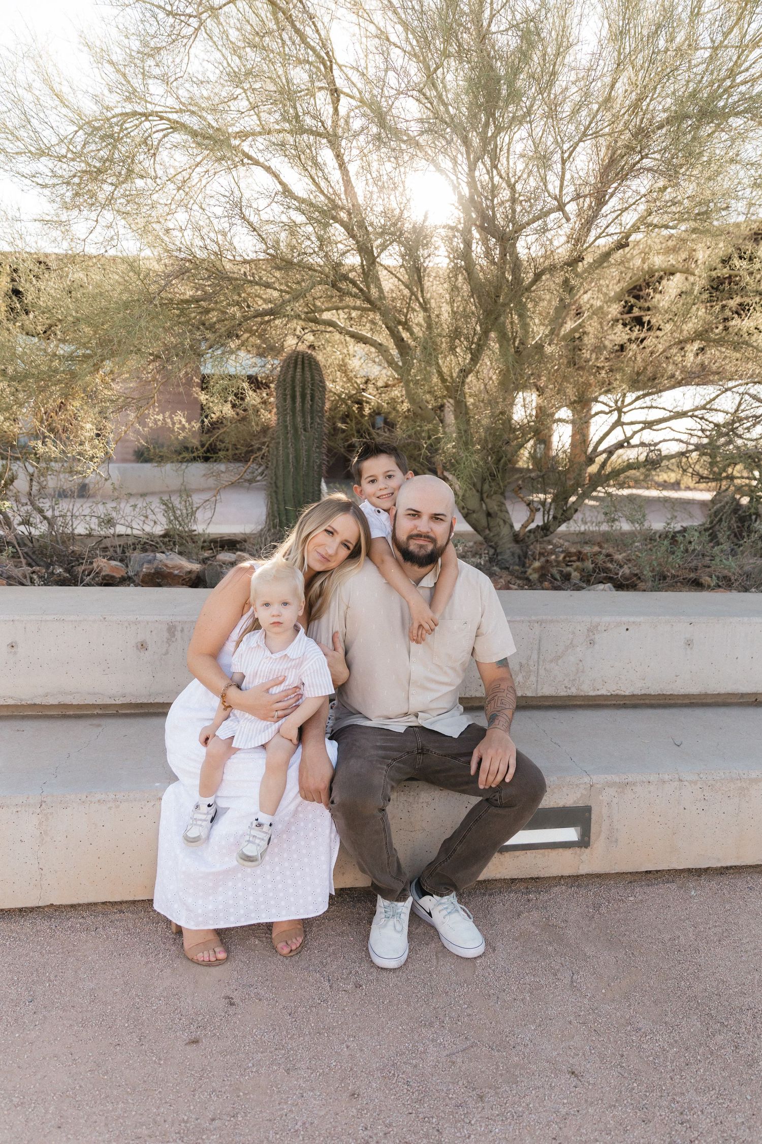 Family in casual white outfits sits together on concrete wall with desert landscape and saguaro cactus in background.