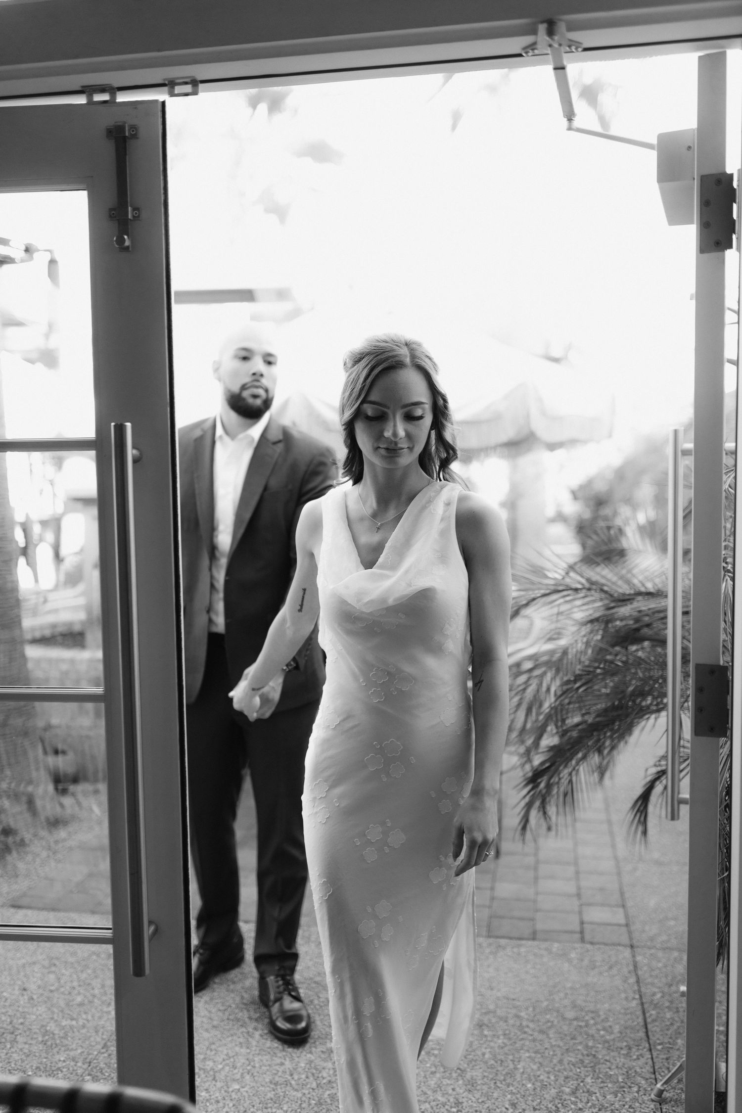 A couple entering through glass doors, with the woman in a white dress leading the way.