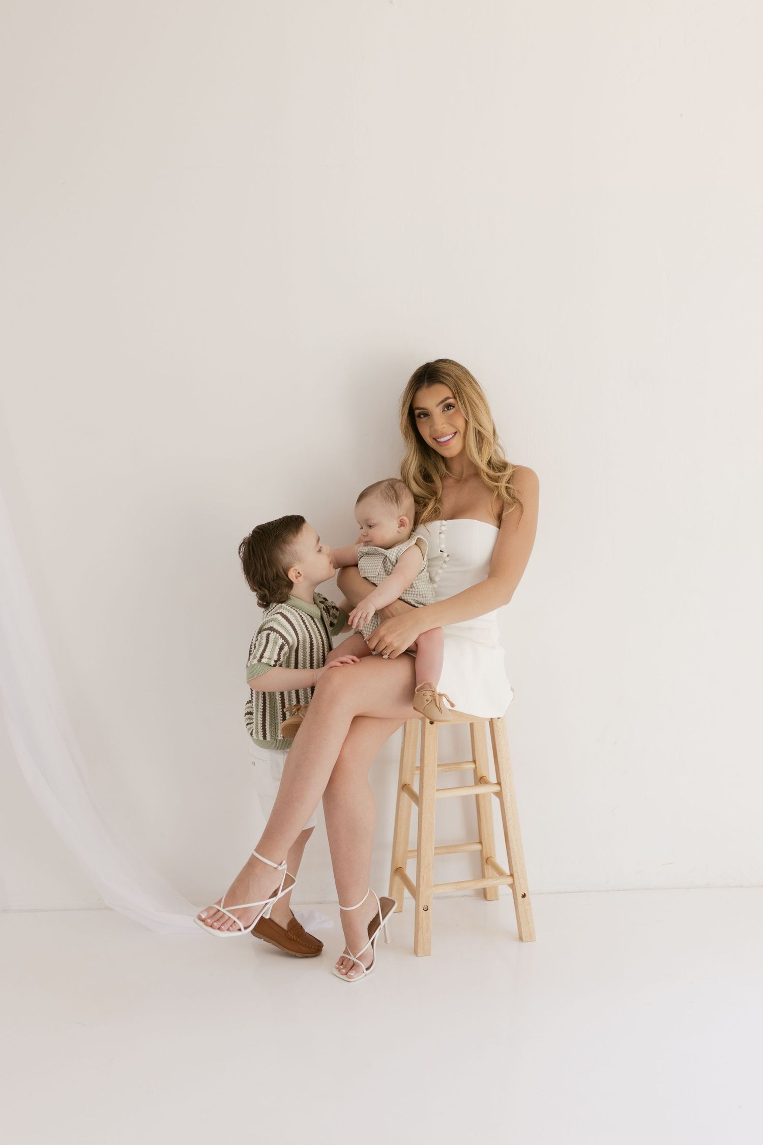 A smiling mother in a white dress sits on a wooden stool holding a baby while a young boy stands beside her.
