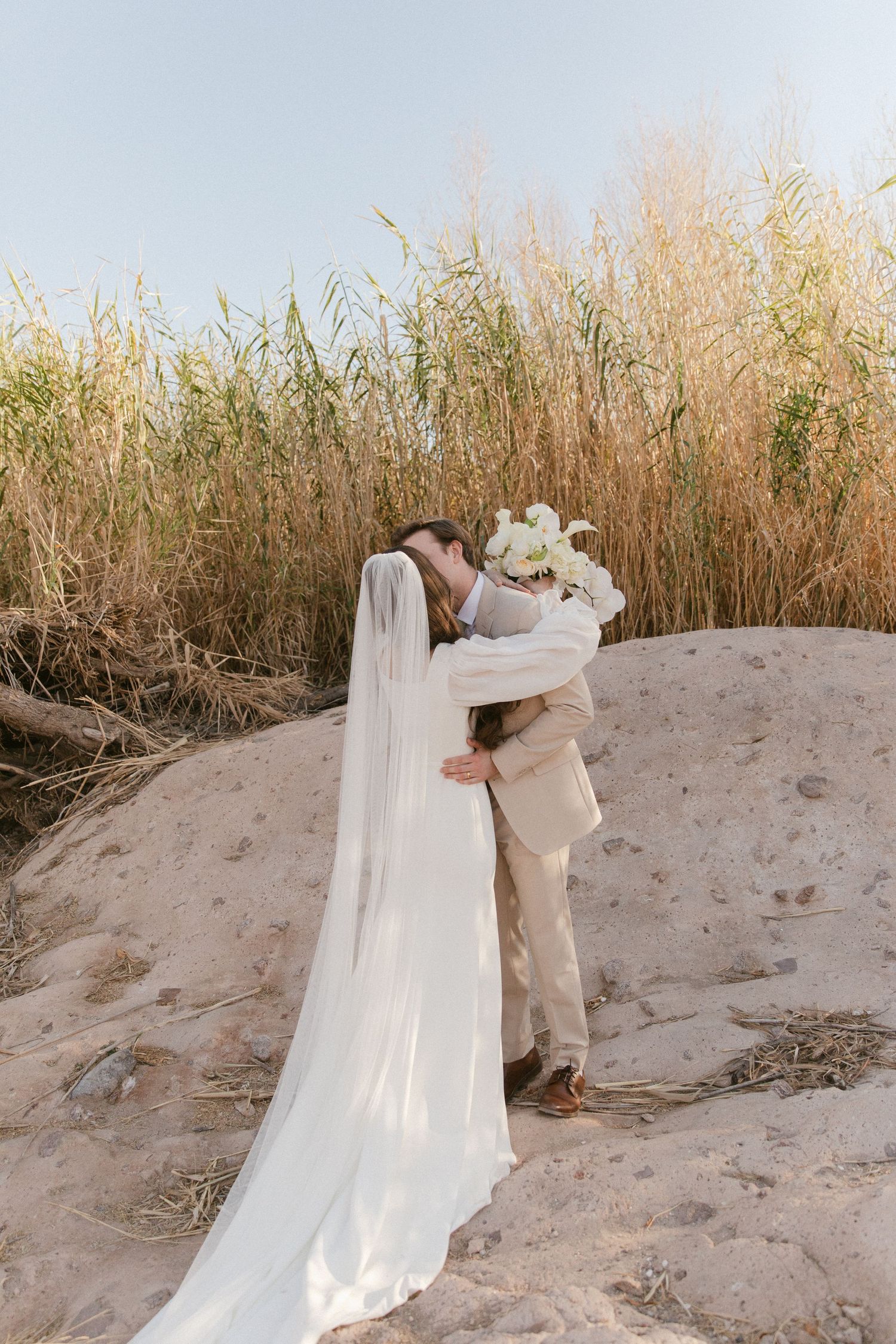 A bride and groom share a kiss on sandy dunes surrounded by tall golden grasses on their wedding day.
