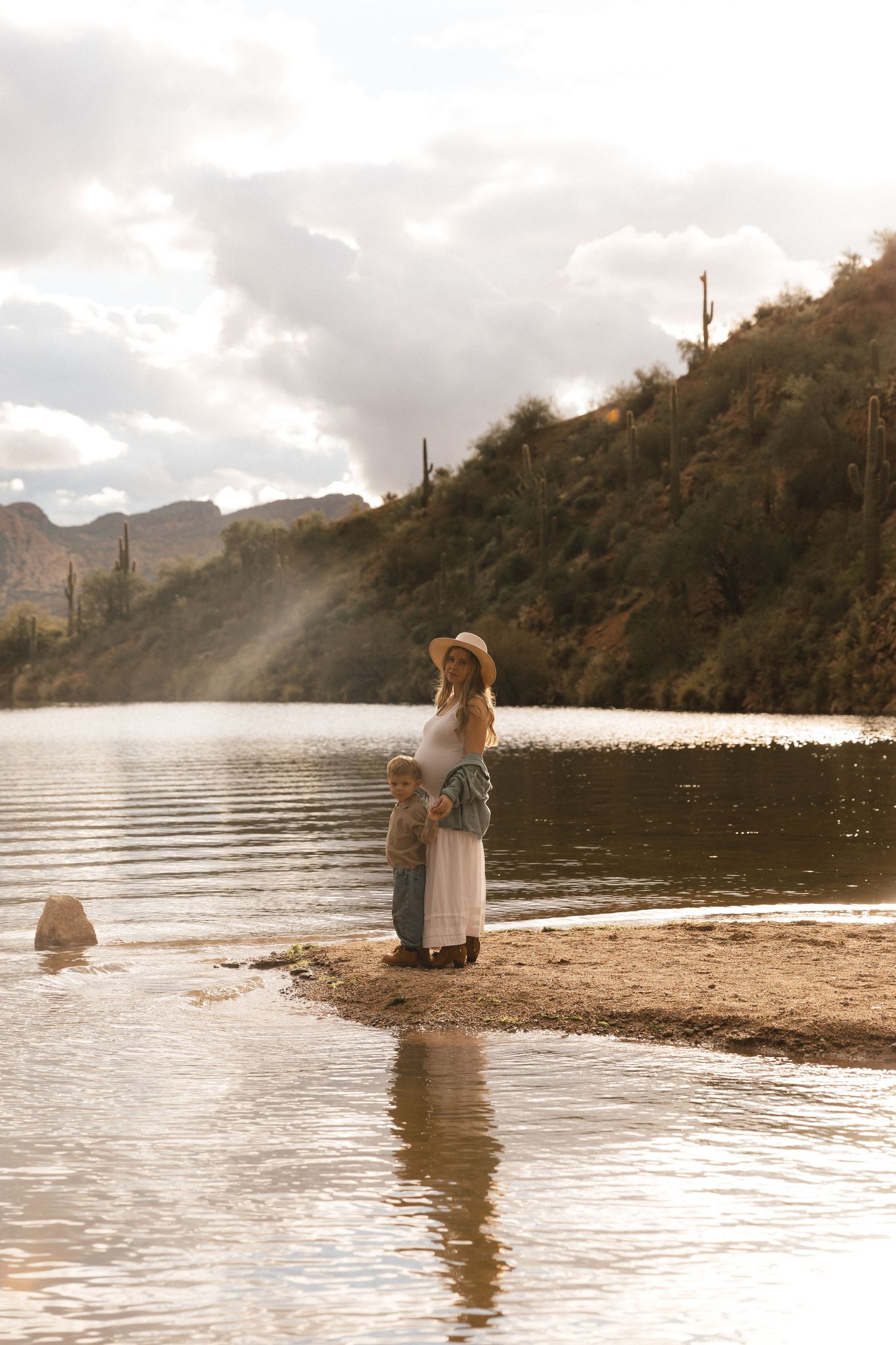 A woman in a hat and white skirt stands with a child by a desert lake, surrounded by saguaro cacti and hills.