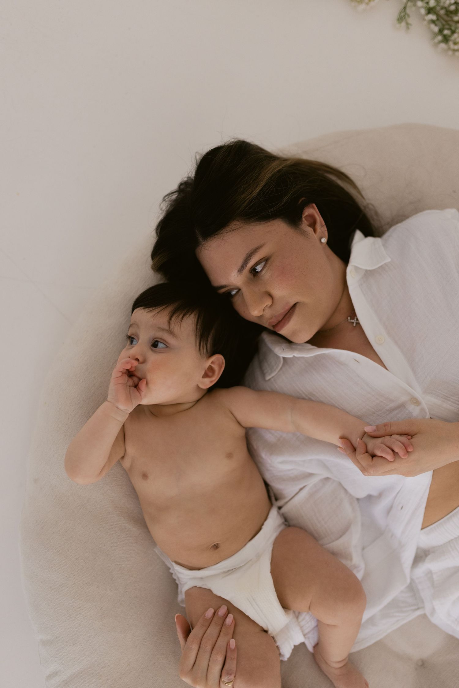 A mother in a white shirt lies beside her baby in a diaper on a soft beige surface, sharing a tender moment.