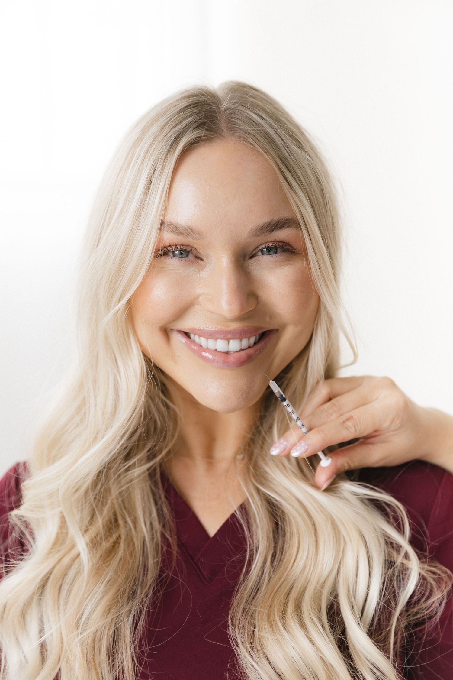 Smiling blonde woman in burgundy scrubs holding a syringe, promoting cosmetic injection services.