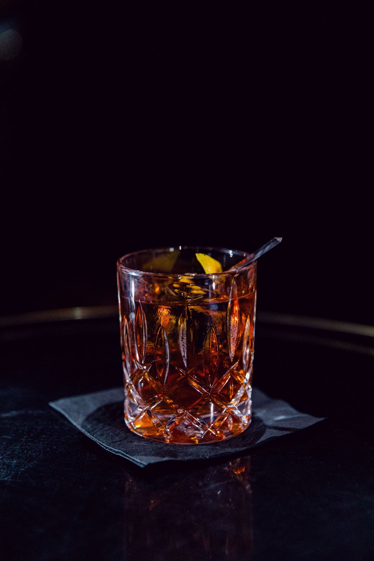 A crystal glass filled with a dark cocktail and garnished with an orange peel against a black background.