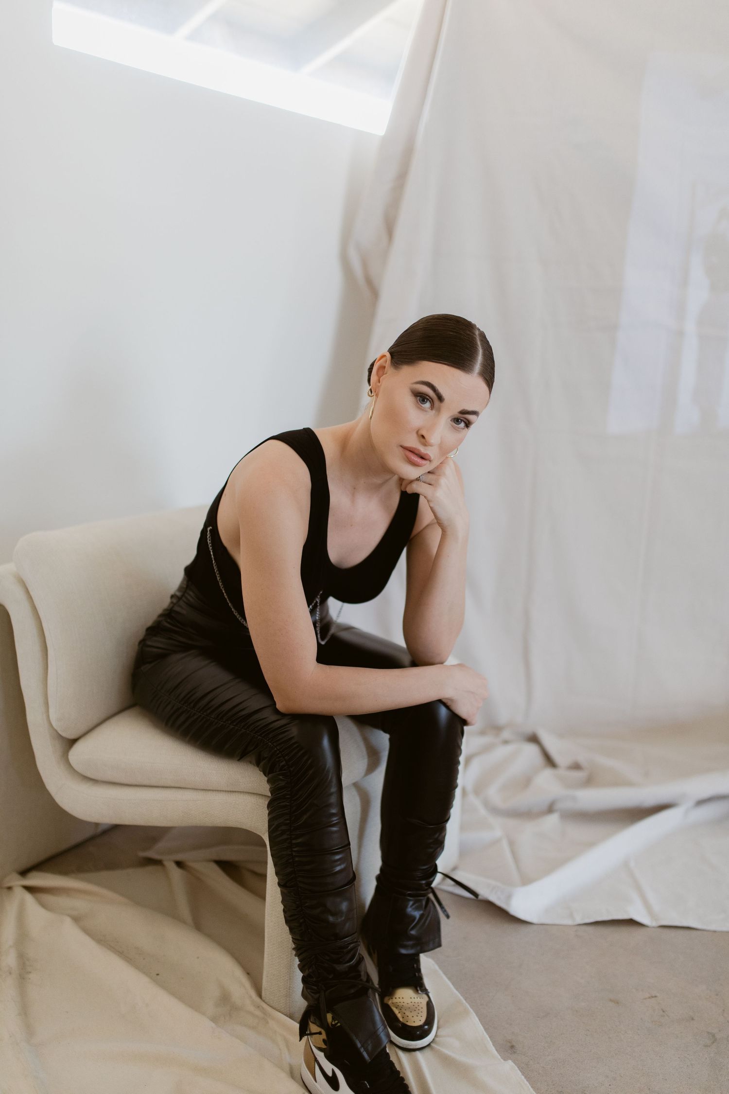 Moody portrait of model in black outfit sitting on beige chair against minimalist backdrop.
