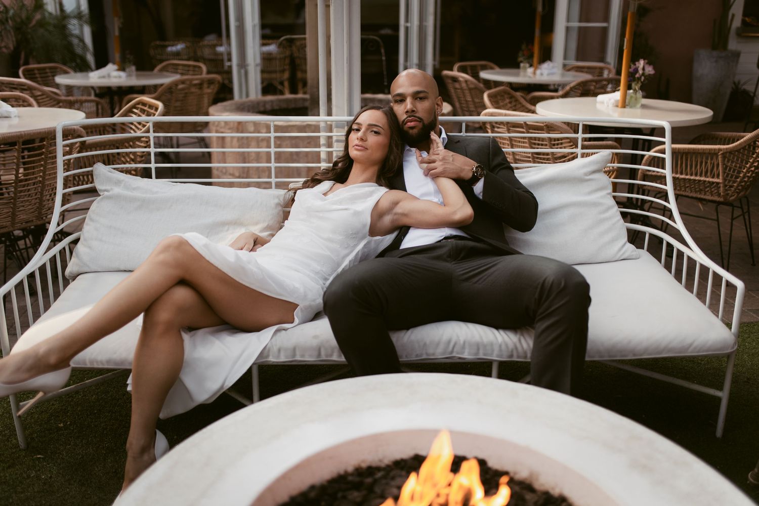 A couple relaxes on outdoor furniture near a fire pit in wedding attire.