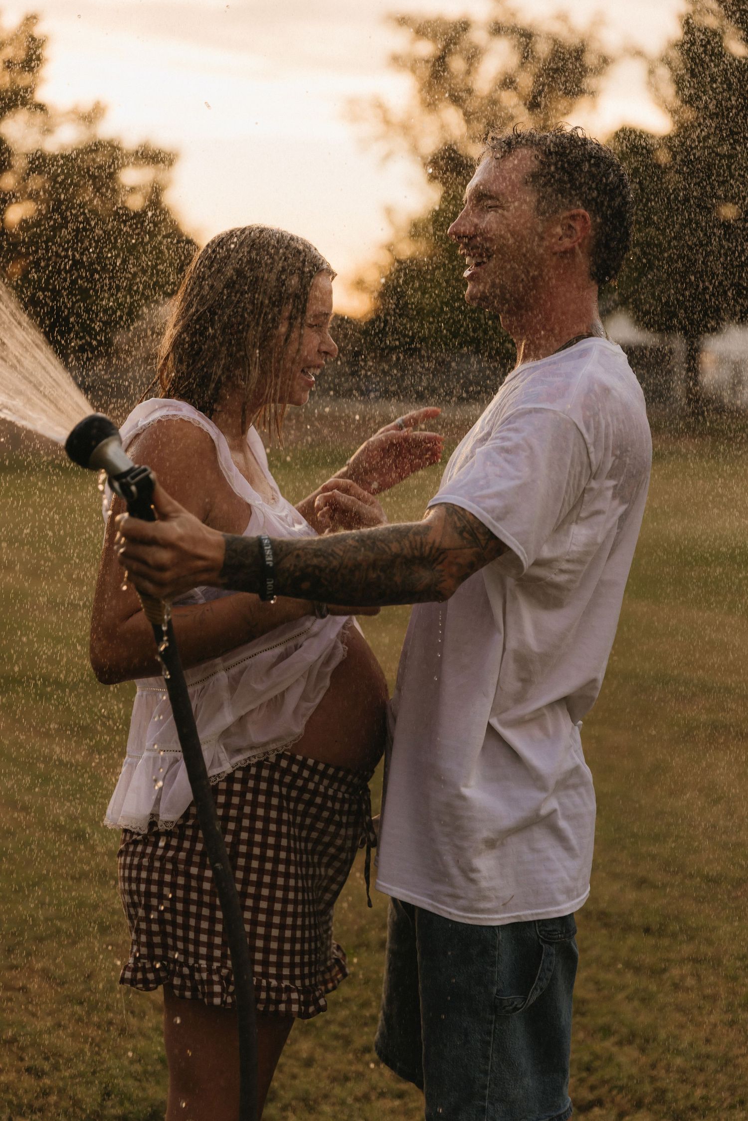 Couple laughing and playing with garden hose at sunset, getting soaked in water spray outdoors.